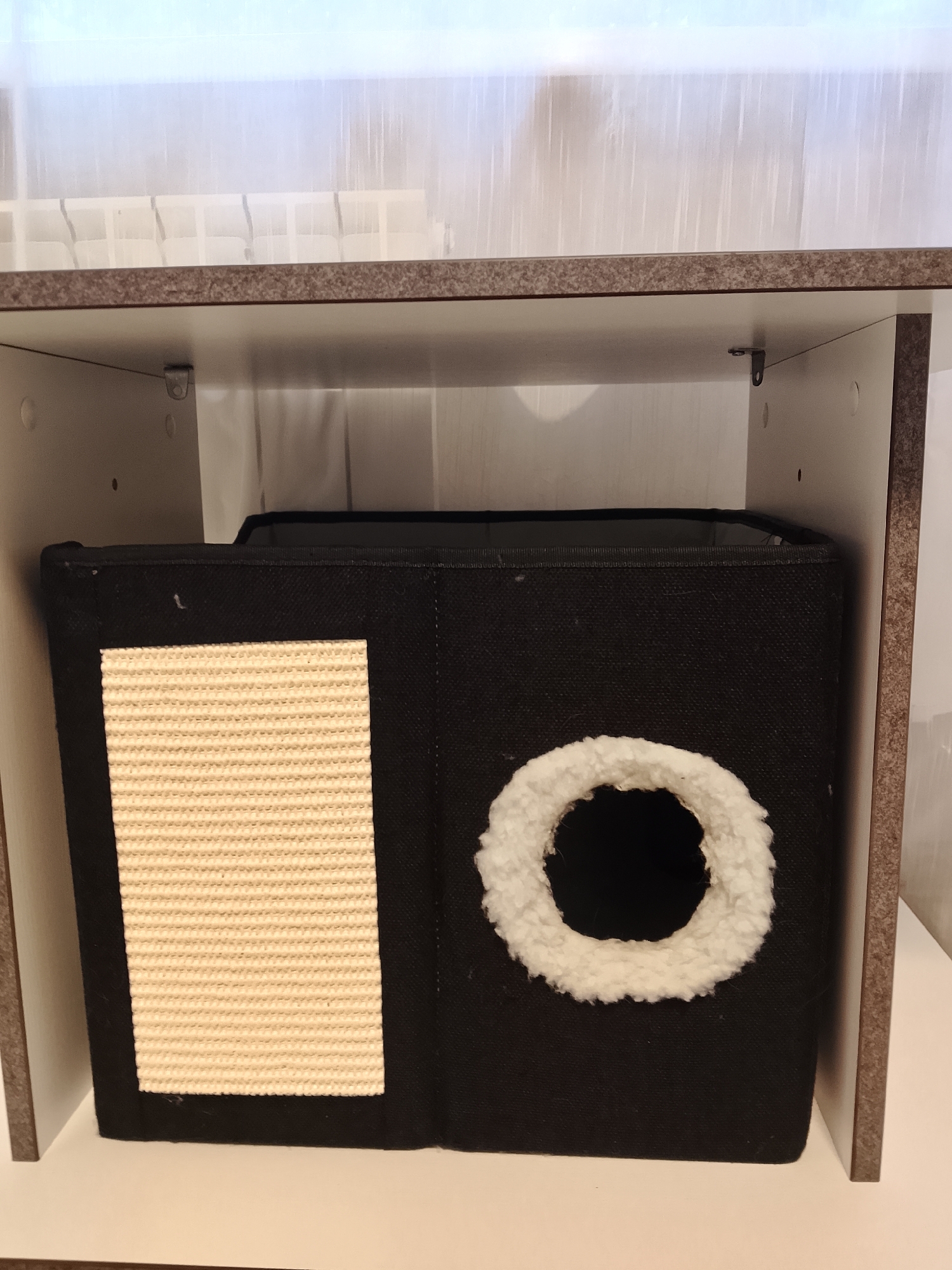 A black fabric cat cube is placed on a white shelf, which is part of a larger shelving unit. The cube has a sisal scratching pad on its left side and a circular entrance hole on its right side, lined with white fluffy material. The background consists of translucent white blinds. The scene appears to be indoors in a domestic setting.
