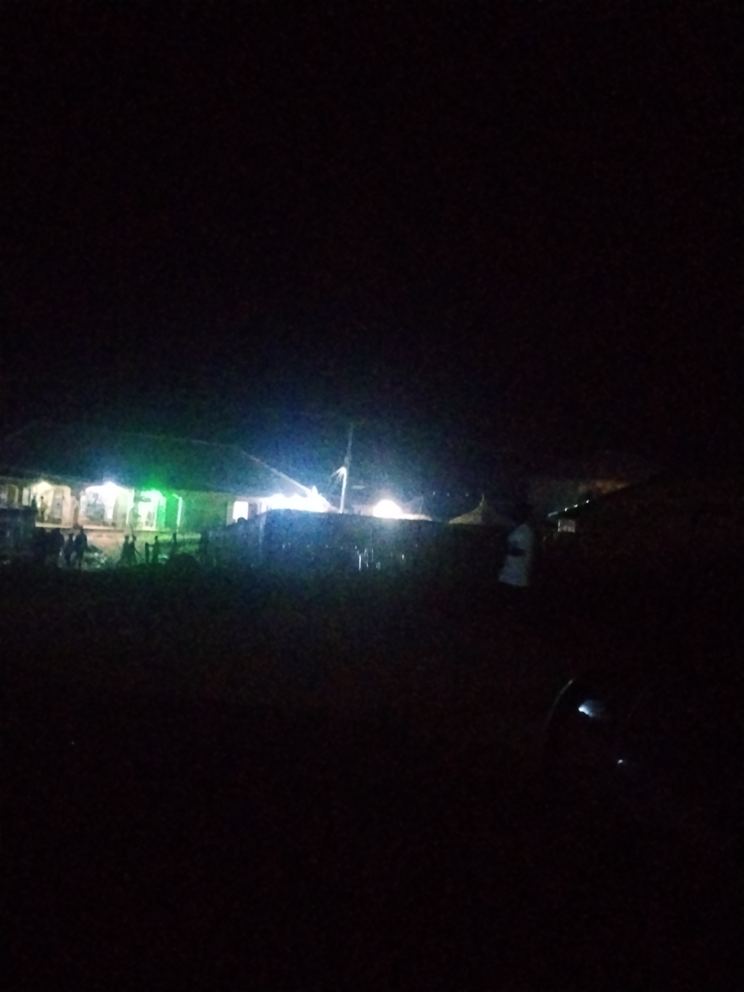 The image depicts a nighttime scene in Durumi, Nigeria. A building with multiple illuminated windows and a green light fixture is visible. Several people are standing and gathered around the building. Some individuals appear to be engaged in activities in front of the structure, and there are also figures further in the background, some of whom are illuminated by lights. The environment is dark, suggesting it is deep night, with artificial lighting from the building and potentially other sources illuminating the immediate vicinity.