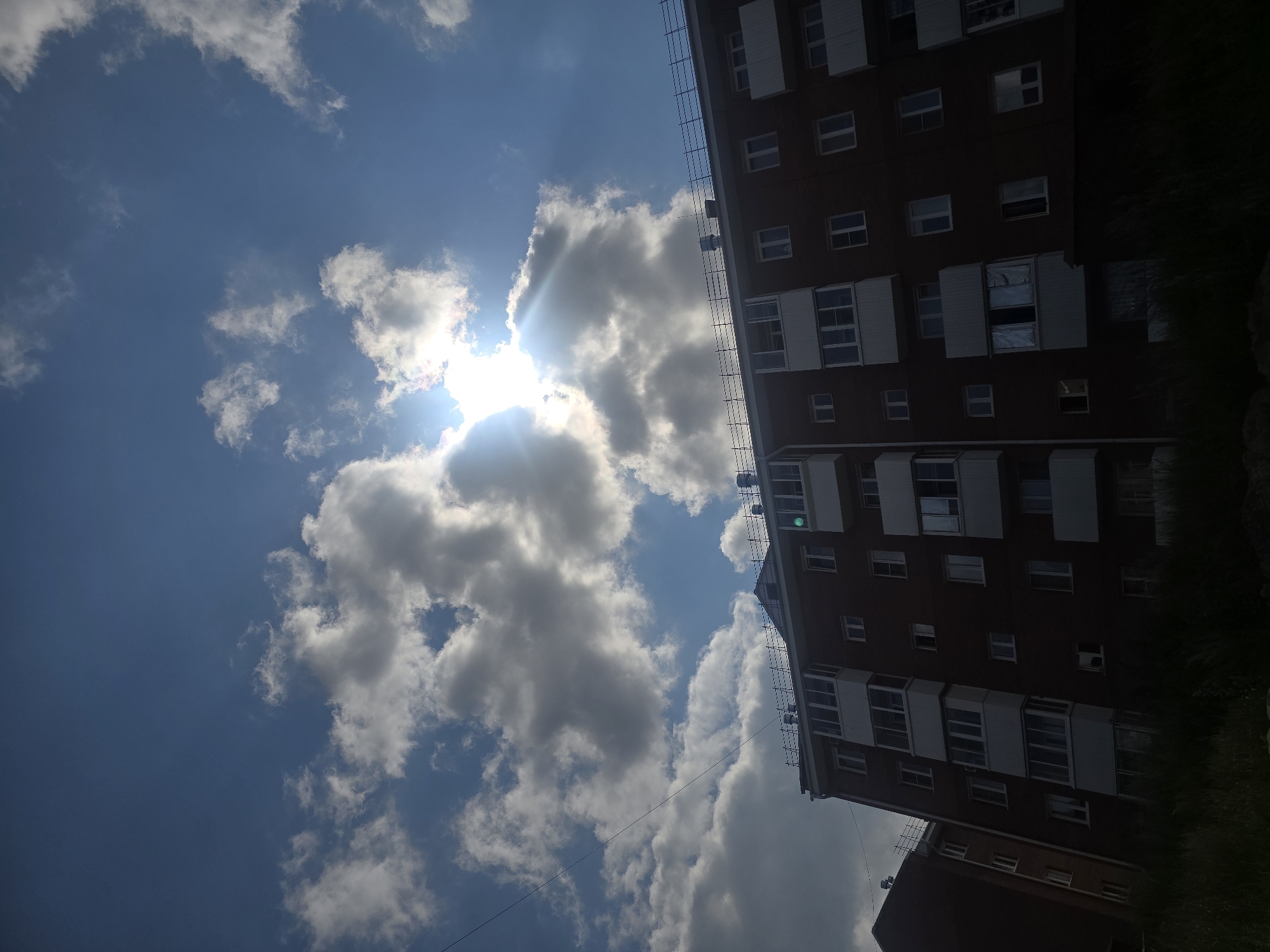 The image shows a residential building in Ulan-Ude, Russia, under a partly cloudy sky. The sun is shining through the clouds, creating bright rays. The building is several stories tall and appears to be made of brick. There are many windows and balconies visible on the facade. The foreground is dark and appears to be overgrown with grass, suggesting a natural or unkempt area. No people are visible in the image.