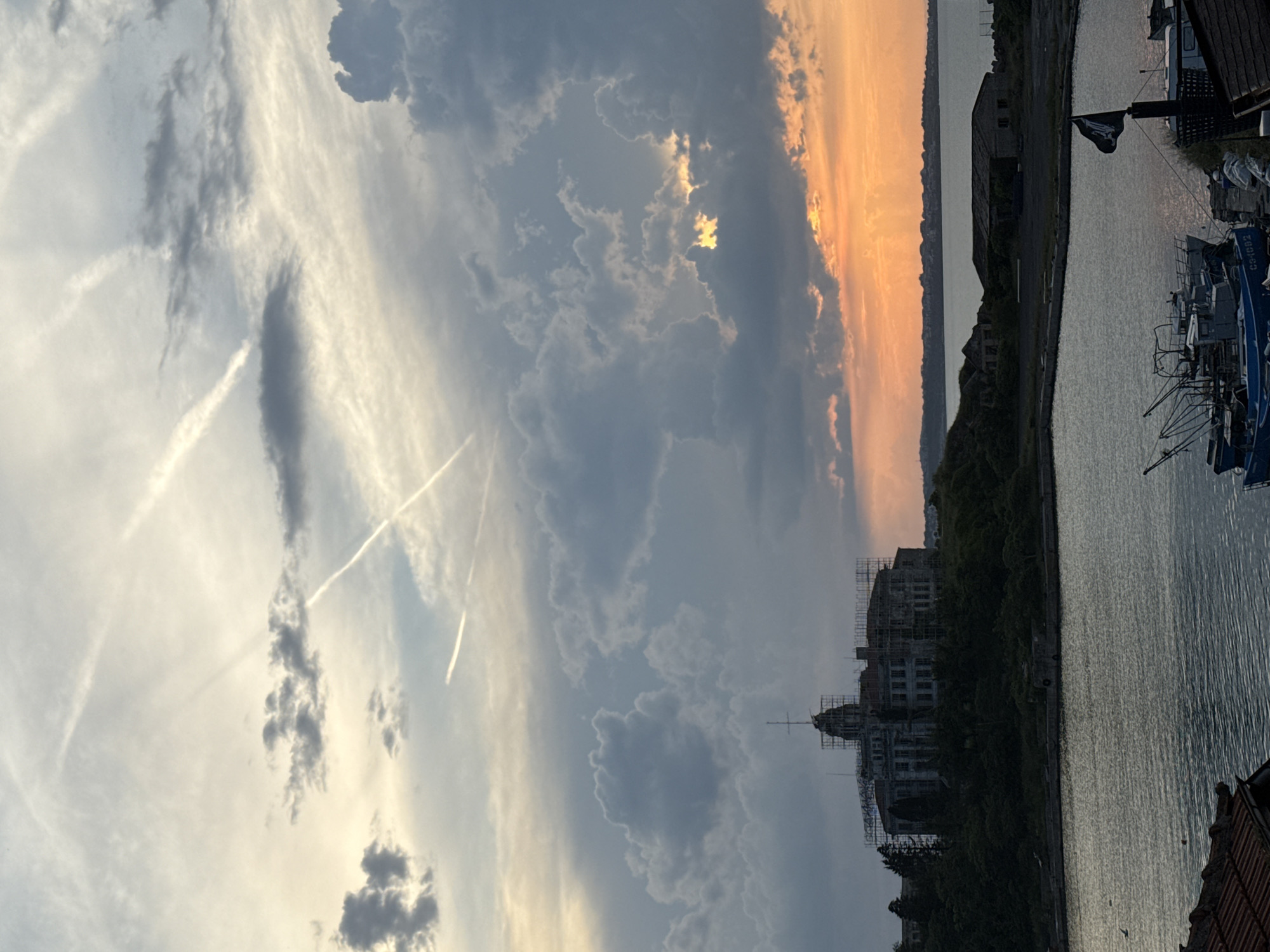 The image captures a picturesque coastal scene at sunset in what is identified as Sozopol, Bulgaria. The sky dominates the upper portion, showcasing a dramatic interplay of clouds. On the left, the sky is a lighter blue-grey with wispy white cirrus clouds and prominent airplane contrails, some forming distinct 'X' shapes. Towards the middle and right, the clouds become darker and more voluminous, dramatically lit by the setting sun which casts vibrant orange and golden hues, breaking through a gap in the clouds and reflecting softly on the water.
The lower half of the image features a calm body of water, likely a sea or a large bay, reflecting the sky's colors and showing gentle ripples. Across the water, a distant landmass or shoreline is silhouetted against the bright horizon. On the right side, closer to the viewer, several structures and boats are visible. A prominent building on the shore appears to be under construction or renovation, enveloped in scaffolding, with a distinct spire or cupola. Parts of other buildings with reddish-brown roofs are also visible in the foreground.
Several boats are moored along the right bank. One blue boat is partially visible, and on its side, the registration "C34092" can be read. A notable detail is a black flag, bearing a white skull and crossbones design (a Jolly Roger), flying from the mast of one of the boats. The overall scene conveys a tranquil evening atmosphere with a blend of natural beauty and human activity along the water's edge.
