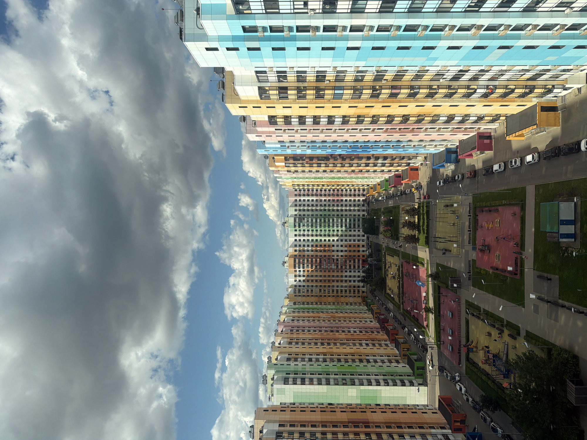 This is a high-angle, medium shot of a residential complex in Moscow, Russia, featuring a row of tall, colorful apartment buildings under a cloudy sky. The buildings are painted in alternating vertical stripes of various bright colors including green, orange, pink, yellow, blue, and light blue. The complex includes several children's playgrounds with red and yellow equipment, and paved courtyards with benches and lampposts. Cars are parked along the streets and in parking lots. The buildings are modern, with numerous windows and balconies. The overall impression is one of a vibrant and densely populated urban area.