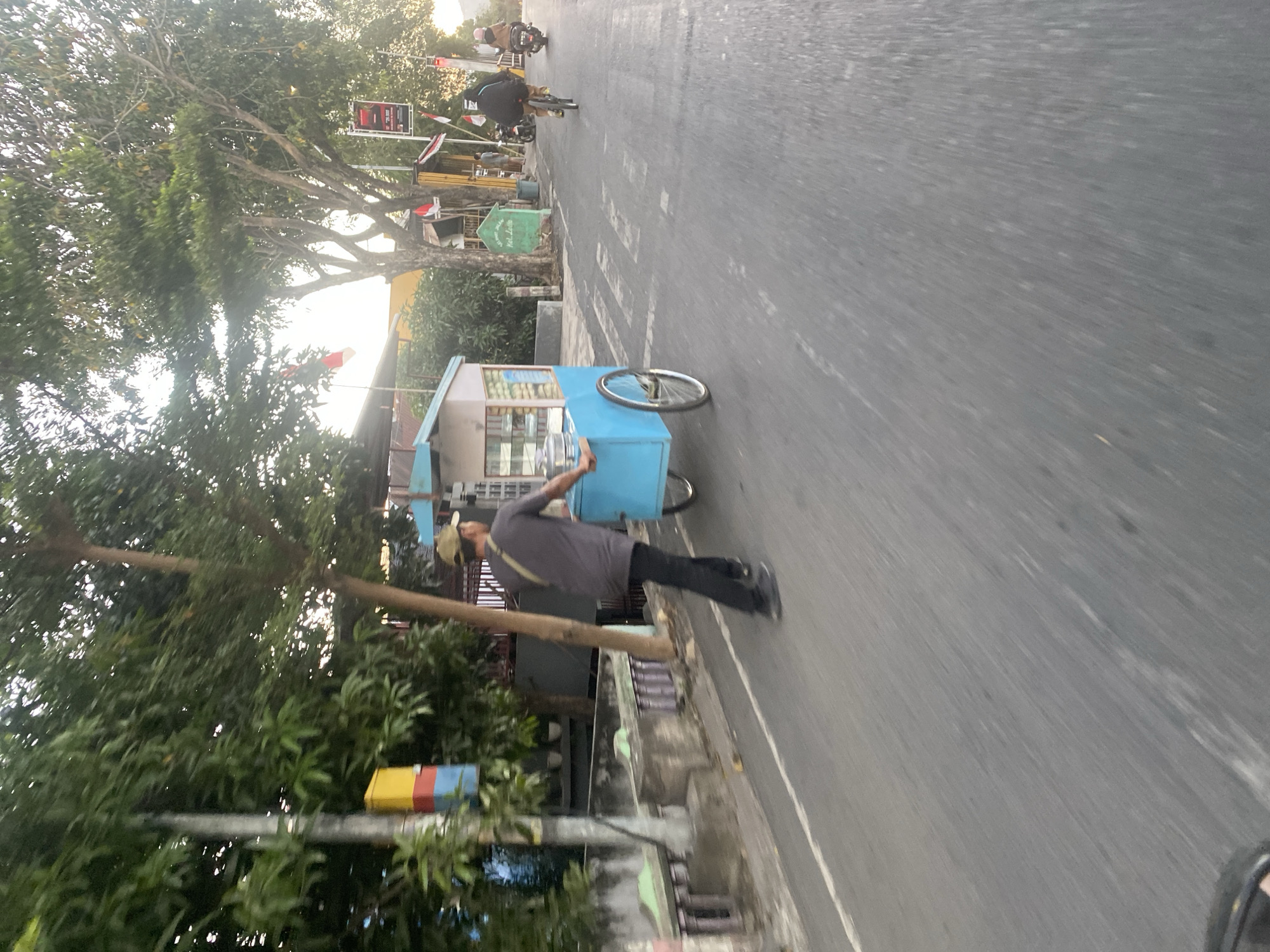 In Bantaeng, Indonesia, a man pushes a blue and white ice cream cart along a paved road. The cart is equipped with a display case showing various items and has two large wheels. The man, wearing a dark gray t-shirt, black pants, and a light-colored baseball cap, is moving from left to right in the frame. The environment features lush green trees lining the left side of the road, with a concrete barrier and some greenery visible behind it. On the right side of the road, there are more trees, some buildings, flags, and a sign that reads "SELAMAT JALAN Kel. Latempa". Two individuals on bicycles and one person on a motorcycle are also present on the road, moving in the same direction as the man with the cart. The lighting suggests it is either early morning or late afternoon, with sunlight filtering through the trees. The road has white lane markings.