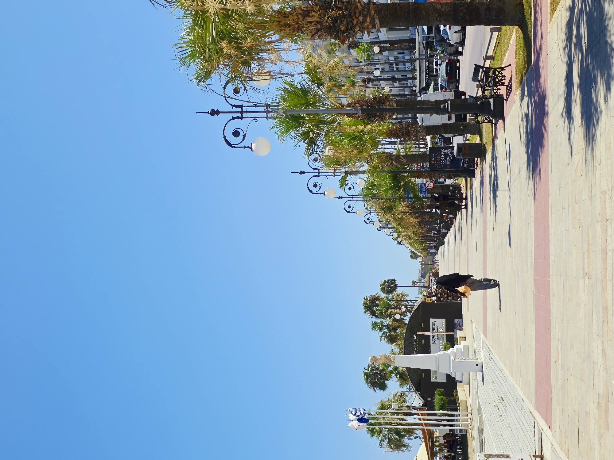 The image depicts a sun-drenched promenade in Larnaca, Cyprus, likely along Finikoudes Beach. A person in dark clothing and a headscarf walks away from the camera on a wide, paved walkway. The walkway is lined with palm trees and ornate lampposts. To the left of the walkway, there are buildings and what appears to be an outdoor seating area for a cafe or restaurant, with umbrellas providing shade. Flags are visible on flagpoles to the left of the walkway, one of which is the Greek flag. In the background, there are buildings, parked cars, and more palm trees, suggesting a coastal urban environment. The sky is a clear, bright blue.
