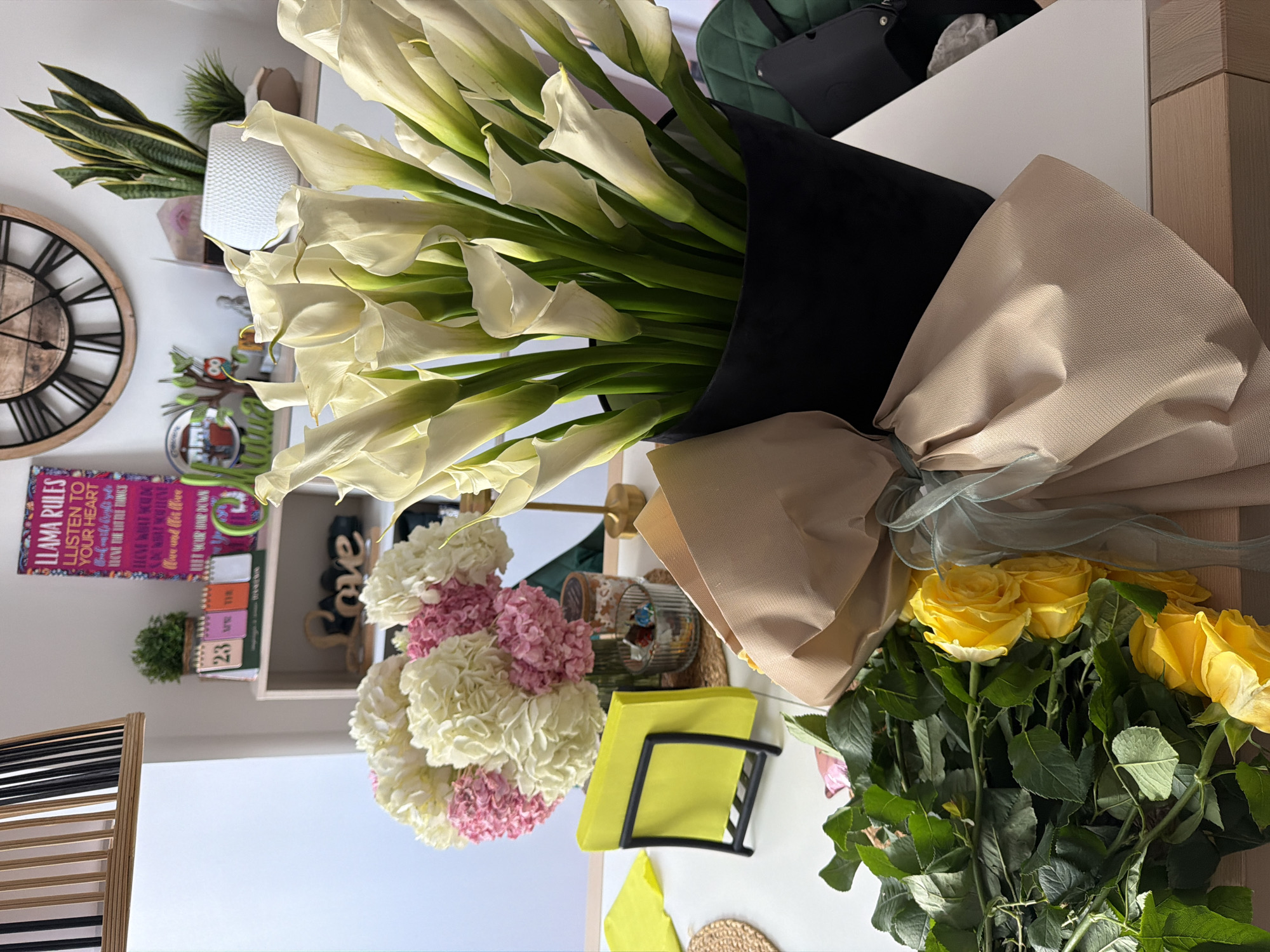 The image shows a bouquet of white calla lilies in a black wrapping with beige fabric accents, alongside a bouquet of yellow roses. There is also a floral arrangement with pink and white hydrangeas. A small glass container holding colorful objects and a black wire holder with yellow sticky notes are on the surface, along with a small woven mat. The setting appears to be indoors, likely a room in a house or apartment. A decorative wall shelf is visible in the background, displaying a clock, plants, and decorative items. A calendar shows the number "23," and a sign with the words "LLAMA RULES" is visible on the shelf. The image does not suggest it is located in Vilafortuny, Spain.