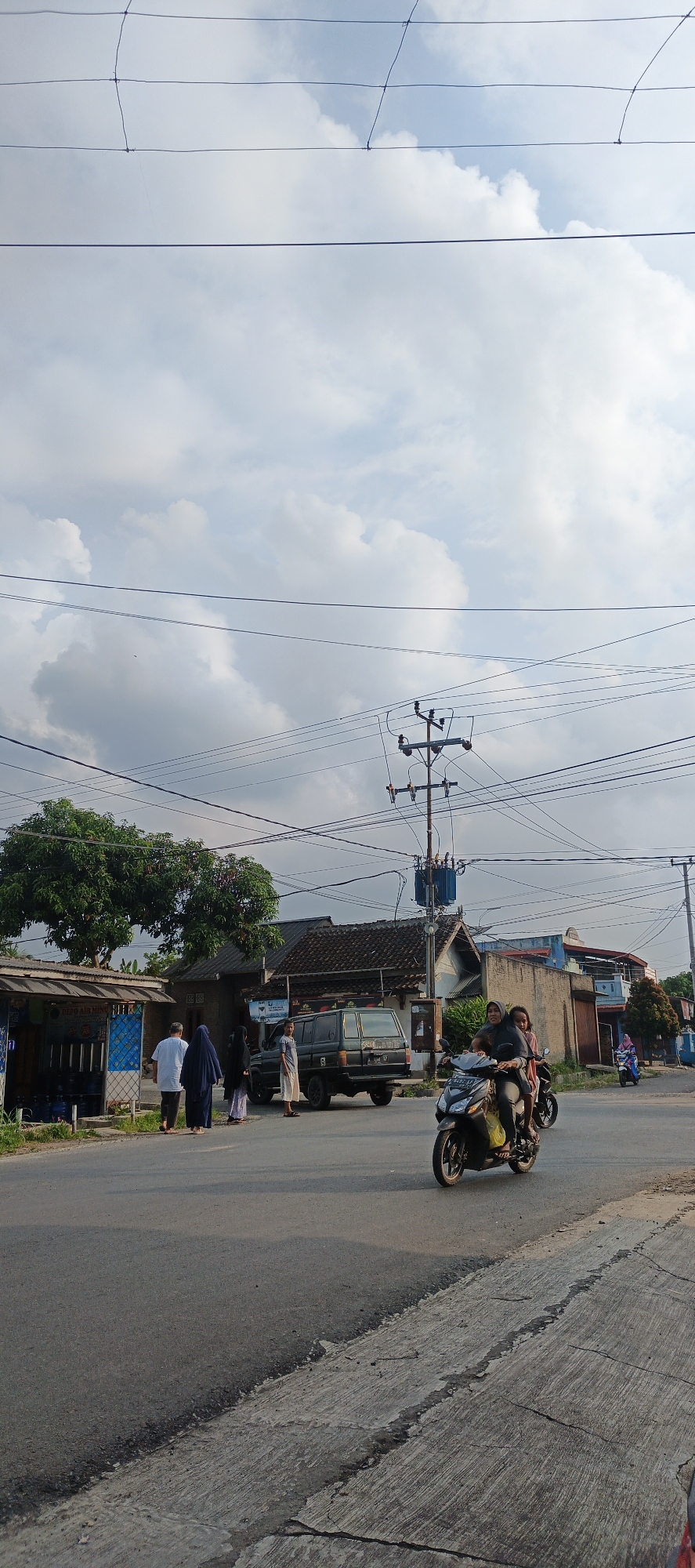 The image depicts a street scene in Sukarame, Indonesia. In the foreground, the asphalt road shows signs of wear and tear with cracked concrete edges. A black motorcycle with two riders is moving away from the viewer. The female rider, wearing a dark hijab, is driving, and a child is seated behind her. Further down the road, a dark green SUV is parked.
On the left side of the street, a small blue and white structure, possibly a shop or kiosk, stands with several bottles visible inside. A group of four people, dressed in traditional Islamic attire, are standing near this structure and the road. Two individuals are facing away from the viewer, while two others are looking towards the parked SUV.
In the background, several houses with dark tiled roofs are visible. A prominent utility pole with transformers and numerous power lines stretches across the sky, which is filled with scattered white clouds against a pale blue backdrop. Trees with dense green foliage are also present. To the right, another motorcycle ridden by a person in a pink headscarf is visible in the distance. The overall lighting suggests it is late afternoon or early morning.