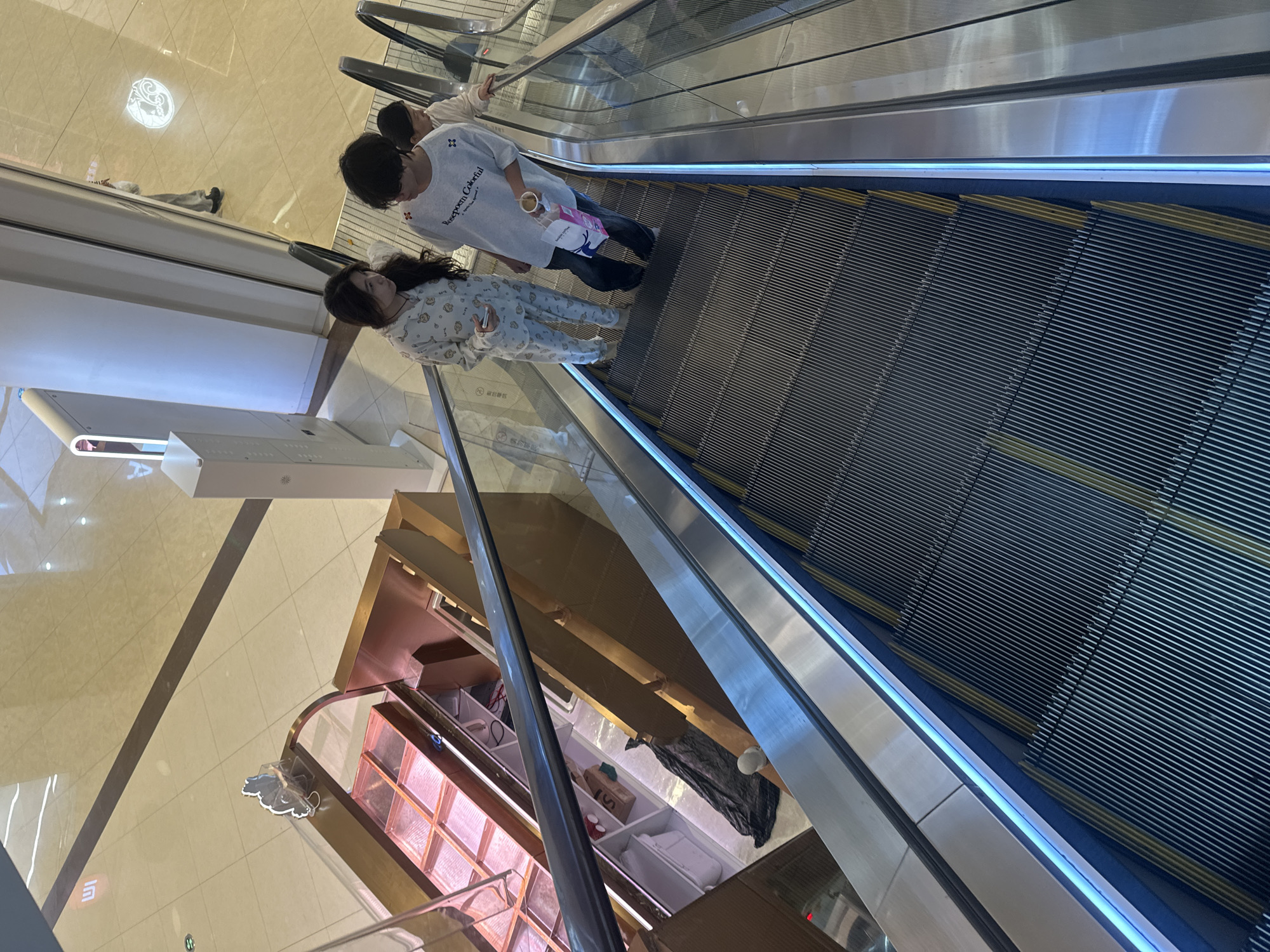 A high-angle shot captures a well-lit shopping mall in Chongqing High-tech Zone, China, featuring a downward-moving escalator. Three individuals are visible on the escalator. A woman in patterned pajamas and a man in a grey graphic t-shirt and dark pants are in the foreground, looking towards each other and engaged in conversation. The man is holding a pink shopping bag and a drink. A third person, possibly a child or a younger individual, is behind them, also on the escalator. To the left of the escalator, a retail display case with rose-gold accents and multiple compartments is visible, suggesting it might be a bakery or confectionery store. The surrounding environment includes a polished tiled floor and modern mall architecture with illuminated signage and pillars. The scene conveys a casual, everyday moment within a commercial setting.