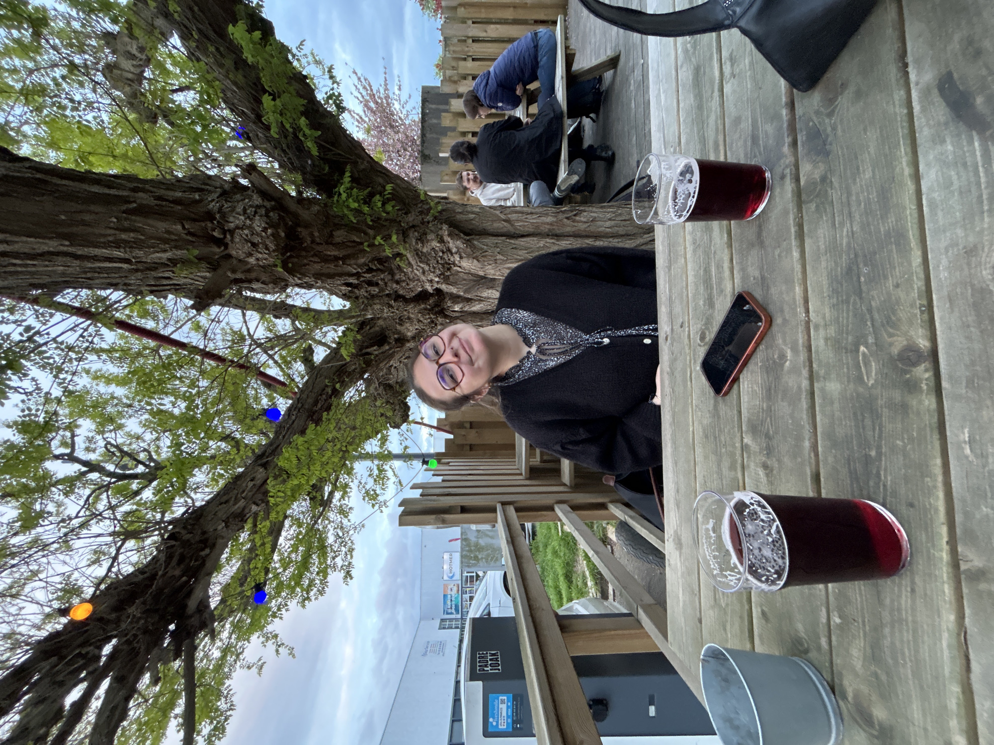 A vertical, full shot shows a woman at a wooden table, with a glass of dark liquid, possibly beer or wine, and a cell phone on the table. The scene is outdoors, with a large tree to the left decorated with multi-colored lights. In the background, other people are sitting at a table. A fence and a building are visible beyond the tables. The building displays the name "PADRE JOAK" and indicates it is for electric vehicle charging. "HOTHER" is also on the building. The location is Villeneuve-d'Ascq, France.