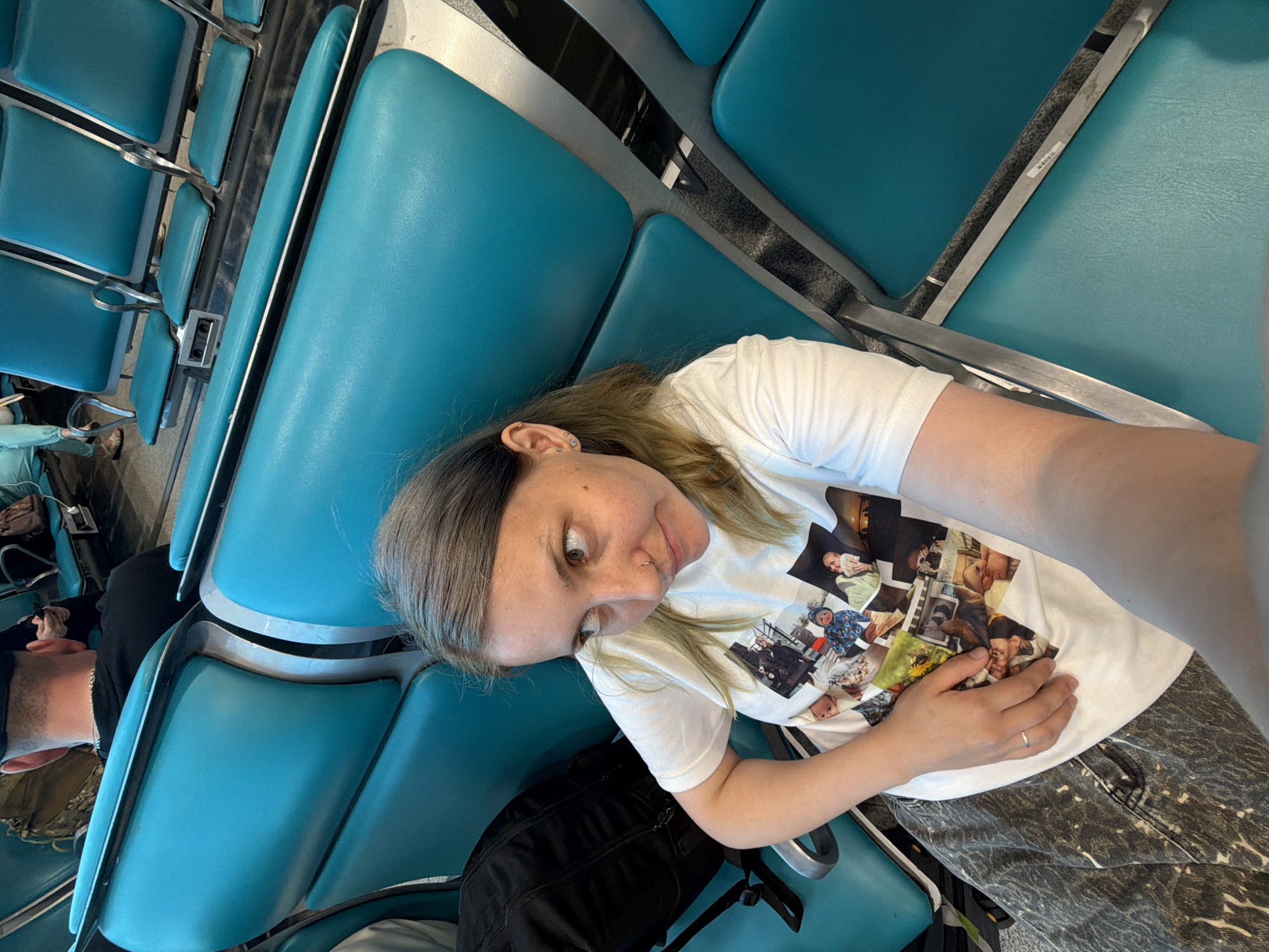 The scene depicts an indoor waiting area in the city of Domodedovo, Russia, characterized by rows of teal-colored public seating with silver-toned metal frames. In the foreground, a female subject, estimated to be in her 20s or 30s, with shoulder-length light brown hair, is captured in a selfie perspective. She has a septum nose piercing and multiple piercings on her left ear. She wears a white short-sleeved t-shirt adorned with a collage of printed photographs, primarily featuring babies and children, and dark, patterned trousers. A silver ring is visible on her left ring finger. Her gaze is directed towards her right (viewer's left). A black backpack rests on the teal seat immediately to her left. In the midground, beyond the primary subject, additional rows of identical teal seats are visible, some equipped with power outlets. In the background, a male individual is seated facing away from the camera, wearing a black t-shirt and a dark baseball cap, with a silver chain around his neck. Another person wearing a light blue garment and sandals is partially visible on the far left. A camouflaged bag is situated behind the seated male figure. The floor appears to be a dark, textured surface.