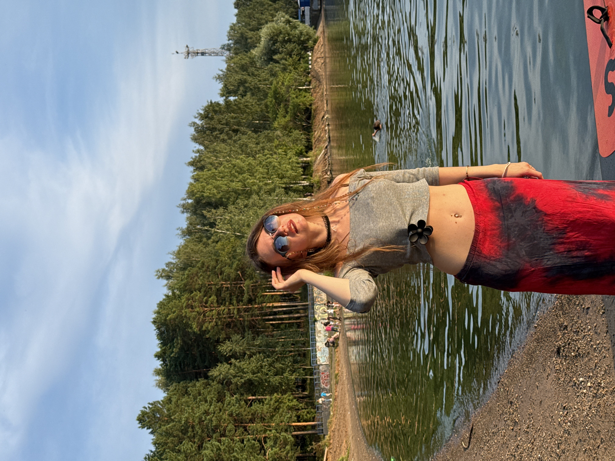 A young woman with long brown hair is standing on a sandy shore, wearing sunglasses, a grey off-the-shoulder top, and a red and black tie-dye skirt. She is posing with her hand on her head and looking up and to the right. The scene takes place outdoors with a body of water in the foreground and a dense forest with a radio tower in the background. The water is calm with ripples, and the sky is partly cloudy. There are a few other people in the background near the water. A bright orange object, possibly a paddleboard, is partially visible in the bottom right corner of the image.