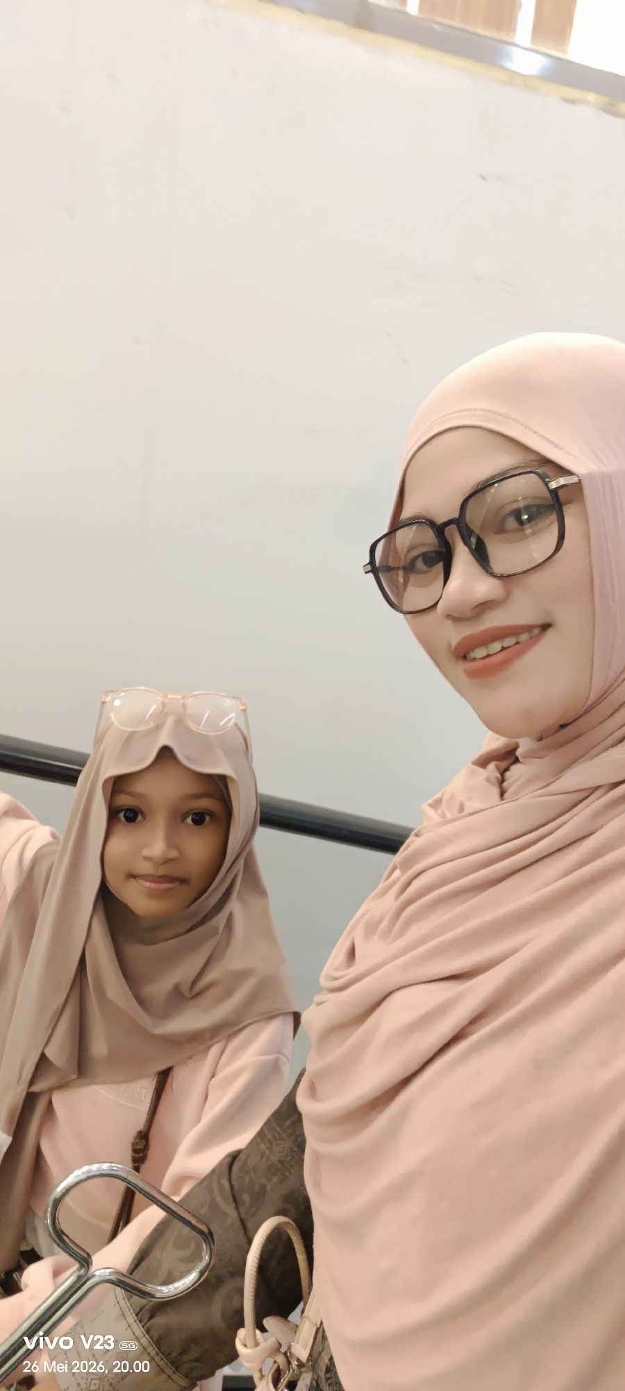 The image captures a selfie of a woman and a girl in what appears to be an indoor location with a plain, light-colored wall in the background. The woman, on the right side of the frame, wears a hijab and black-framed glasses. She has light skin, dark eyes, and is smiling slightly, revealing a reddish-brown lipstick. The girl, on the left side, is also wearing a hijab and has a pair of glasses perched on top of her headscarf. She has dark eyes and light skin, and she is looking directly at the camera with a neutral expression. A black railing is visible behind them, suggesting they may be on an escalator or near a staircase. In the foreground, part of a purse or bag with a silver handle is visible. The image has a timestamp "vivo V23 26 Mei 2026, 20.00". It is unknown if the location is Lhokseumawe, Indonesia.