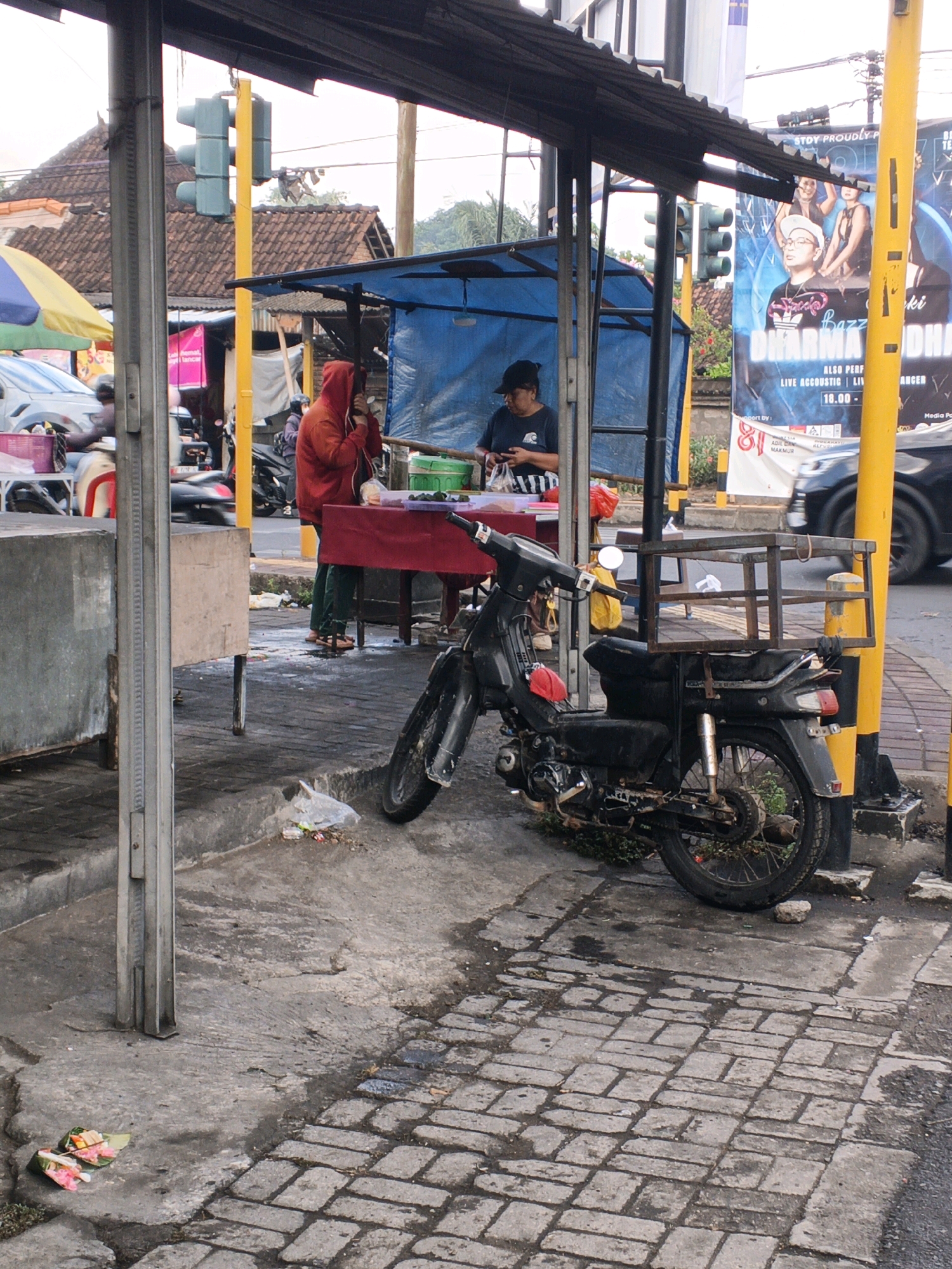 A street scene in Denpasar, Indonesia, features a makeshift stall with a blue tarp roof, displaying goods on a red tablecloth. Two individuals are present: one wearing a red hooded sweatshirt and green pants, holding a white object to their face, and another wearing a dark t-shirt and black cap, handling what appears to be food items. A black motorcycle, equipped with a metal rack on the back, is parked next to the stall. The ground is a mix of concrete and brick paving. Traffic lights and billboards are visible in the background, indicating an urban setting. Debris, including plastic bags and food scraps, litters the ground around the stall and motorcycle.