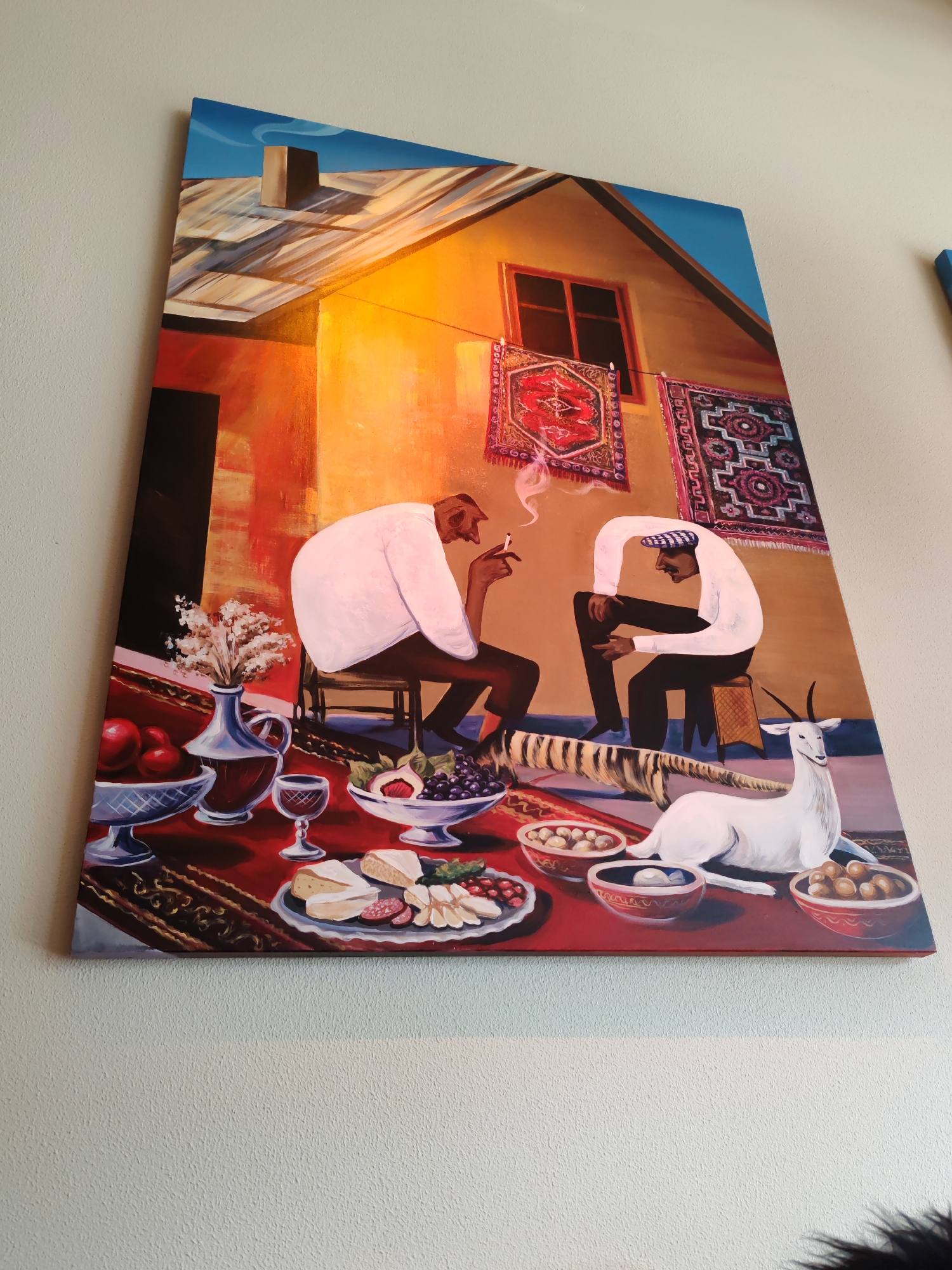 A painting on a wall depicts two men and a goat. The painting is a tall, rectangular canvas. The scene shows a bright orange building with a brown roof and a blue sky. Two rugs are hanging on the wall of the building. In the foreground, there is a table with food and drinks. There are bowls of fruit, cheese, and other items on the table. Two men are sitting at the table, and a white goat is lying down next to them. The painting is in a room with a white textured wall. The location is not specified to be in Syktyvkar, Russia.