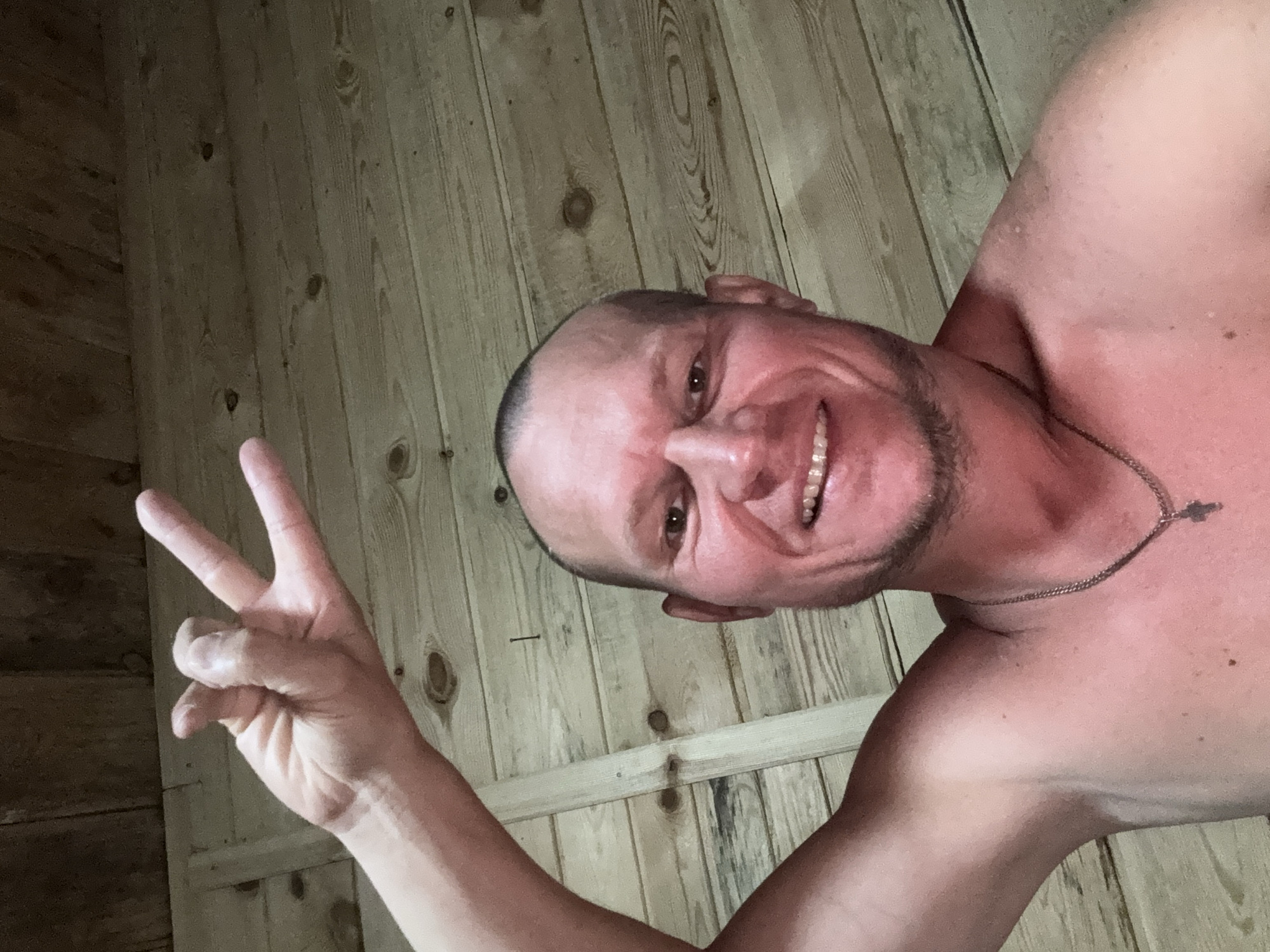 A shirtless man with a shaved head and a beard smiles at the camera while holding up his left hand in a peace sign. He is wearing a silver chain with a cross pendant. The background is a wooden wall. The image was taken in Zharbulak, Kazakhstan.