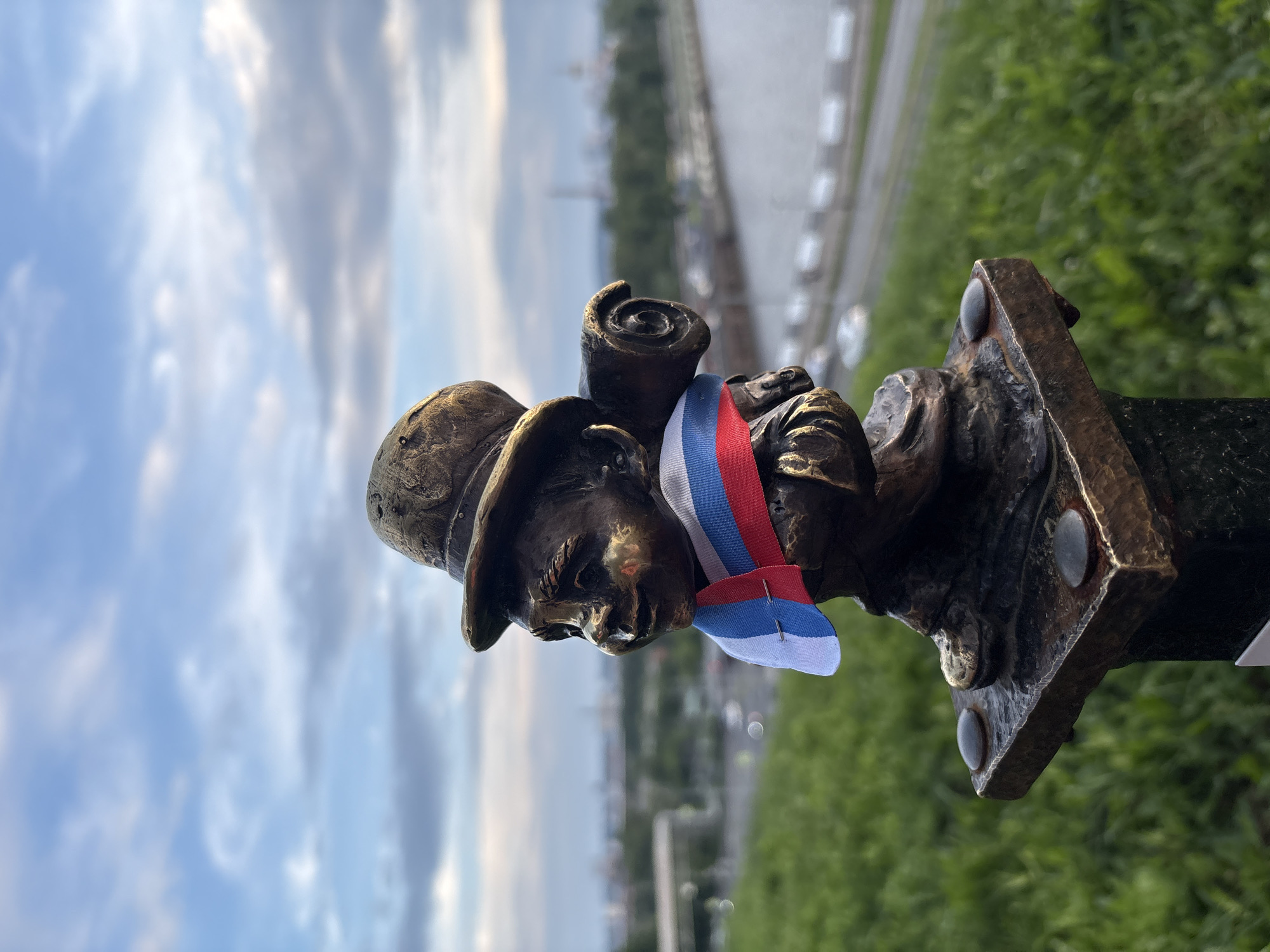 A bronze statue of a dwarf wearing a hat and a sash in the colors of the Russian flag stands on a pedestal outdoors. The dwarf is carrying a rolled-up object on its back. The statue is positioned on a grassy hill overlooking a river and a city in the background. The sky is cloudy. This is the statue of "Nikolai," a dwarf commemorating Nikolai Alexandrovich, located in Izhevsk, Russia, on the embankment of the Izh River.