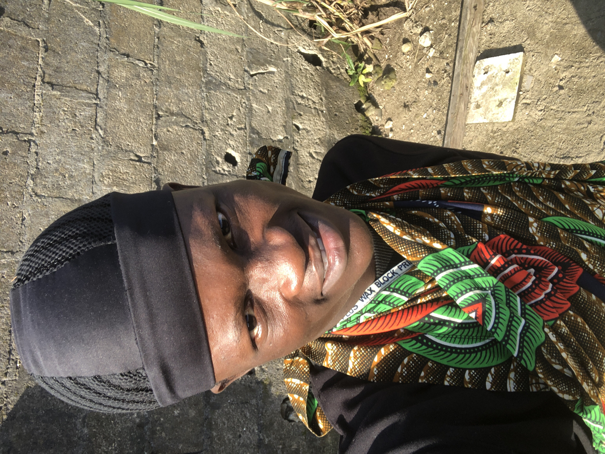 A medium shot, eye-level, outdoor selfie shows a young Black male facing forward and slightly to the right, looking at the camera. He is wearing a black skullcap with a mesh top and a black long-sleeved shirt. A colorful, patterned fabric is draped around his neck and chest, featuring red, green, and brown designs. The background consists of a rough, textured concrete block wall that is partially shadowed, with dry grass and dirt on the ground. A sliver of green grass is visible on the right side of the frame. The man has a slight smile, and his skin is smooth with a warm tone. The scene is illuminated by bright sunlight, creating strong highlights on his face and casting shadows on the wall. The ground appears to be a dirt path or uneven terrain with scattered small rocks and a piece of concrete debris. The overall impression is one of a candid, sunny day in an urban or semi-urban environment, likely Ado, Nigeria.