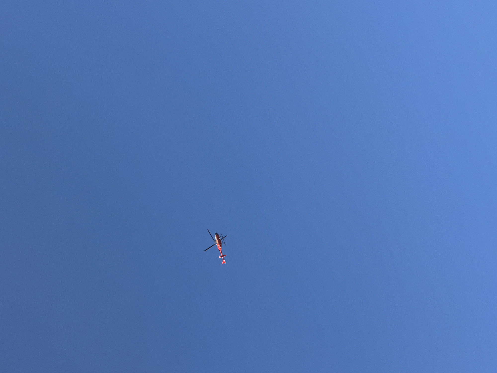 A red helicopter with black rotors and a black tail rotor is captured in flight against a clear blue sky. The helicopter is positioned in the upper left quadrant of the image. The background is entirely composed of the vast expanse of the sky, with no visible ground features, buildings, or other objects.