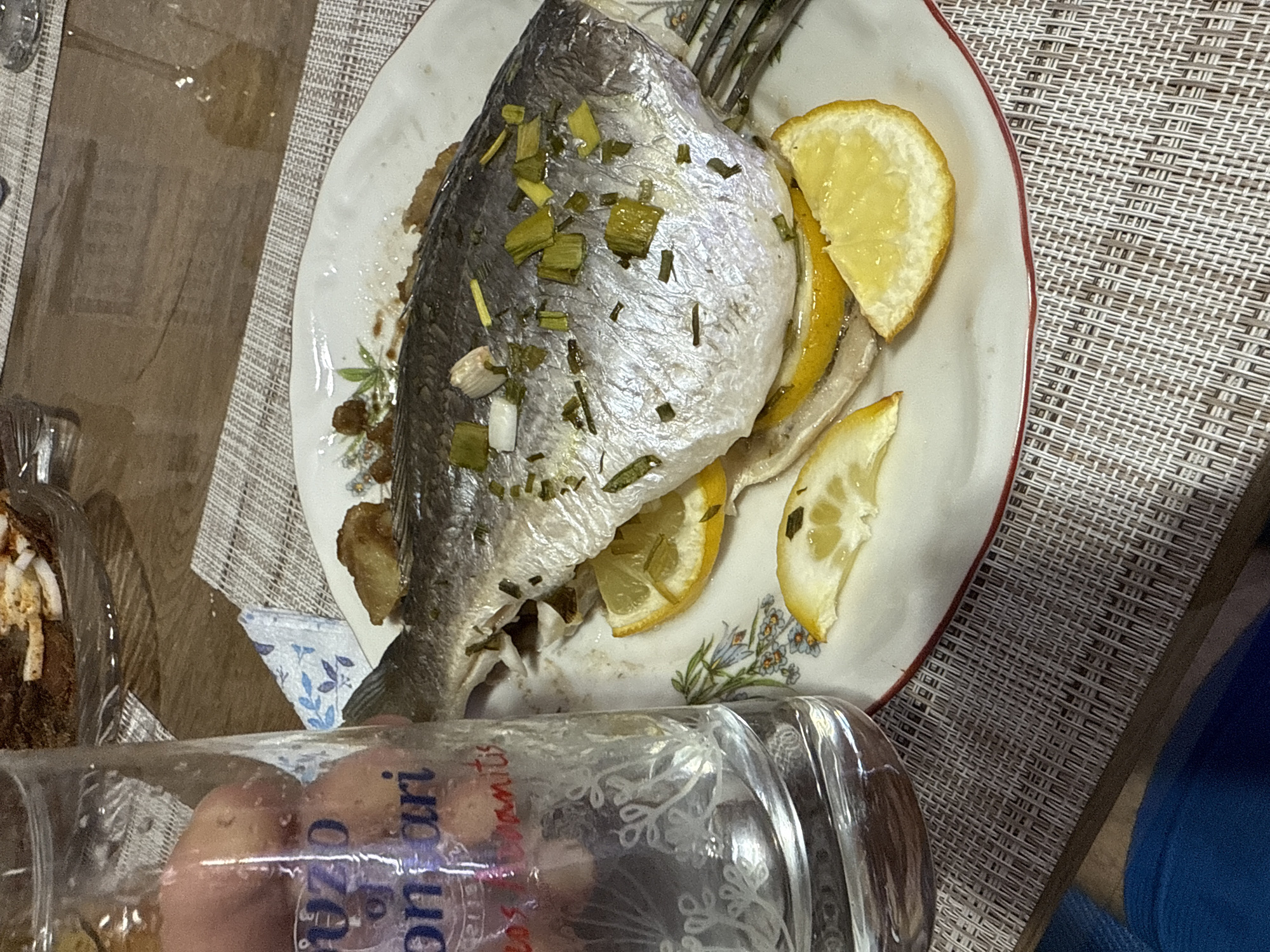 The image shows a plate of cooked fish garnished with lemon slices and green herbs, placed on a woven placemat. A glass, partially filled with an unidentifiable liquid, is visible in the foreground, obscuring part of the plate. Another dish or container is partially visible to the left. The background is a blurred interior setting. The location in the city of Sofia, Bulgaria is not identifiable from this image.