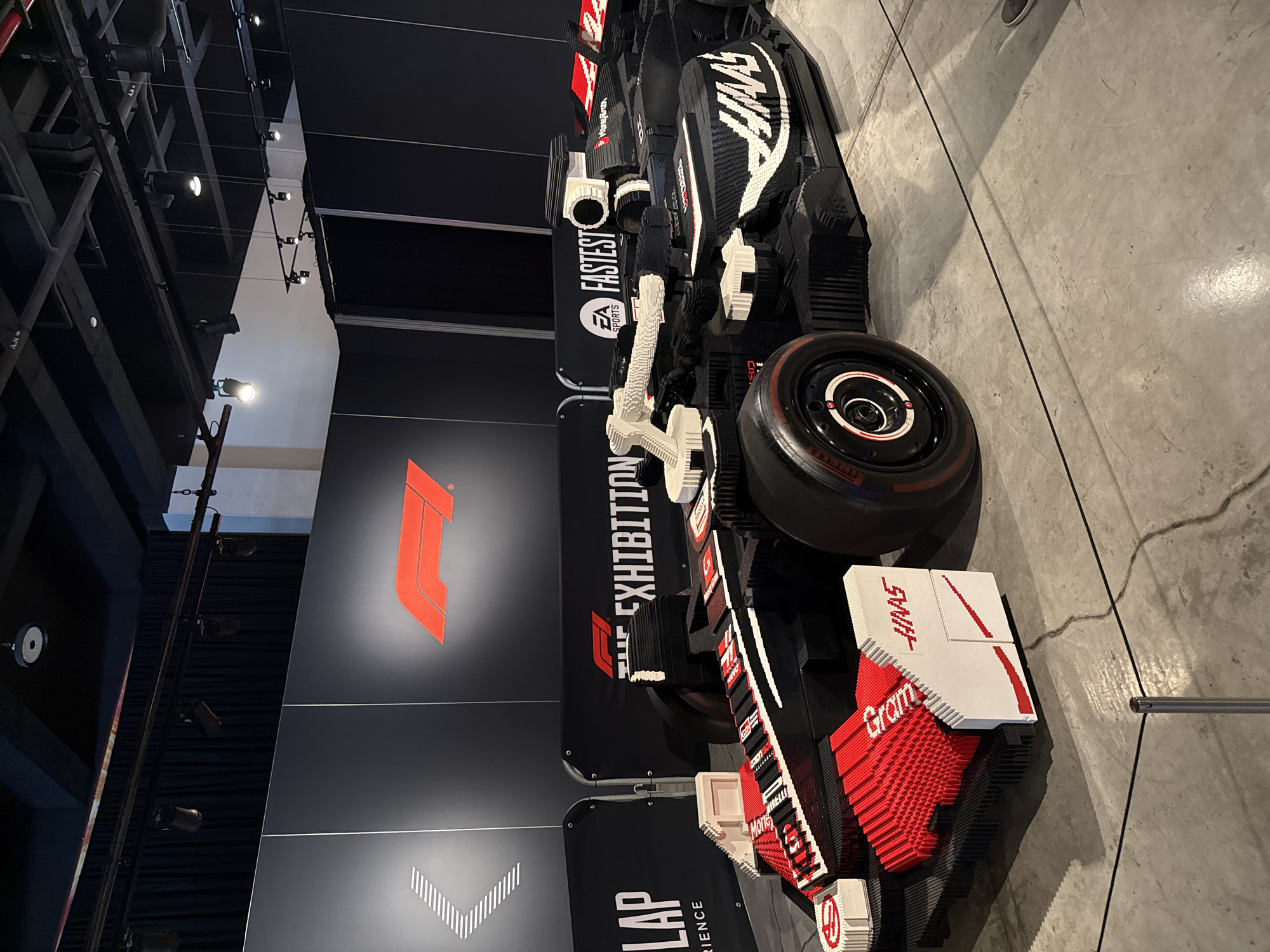 A photograph taken indoors depicts a model of a Formula One race car constructed from Lego bricks. The model is positioned alongside a wall which features the Formula One logo. Adjacent to the logo, the word "EXHIBITION" is vertically oriented. The model features the "HAAS" logo. The flooring appears to be concrete. The location is in Mexico City, Mexico.