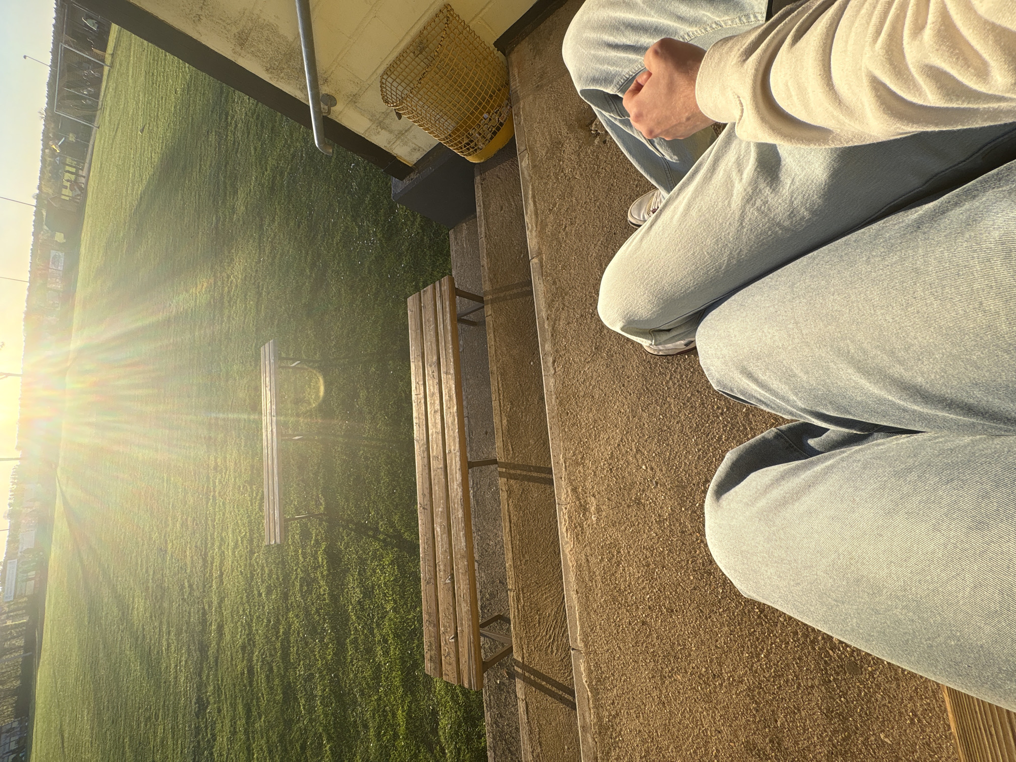 The image shows a person sitting on concrete steps next to a wooden bench, with a grassy field extending into the distance. The person is wearing light-wash denim jeans, a white sweater, and white sneakers. A yellow trash can sits near the top of the steps. The bench is adjacent to the grassy field, and the scene is bathed in bright sunlight. Based on the location, this photo may have been taken in Wegberg, Germany.