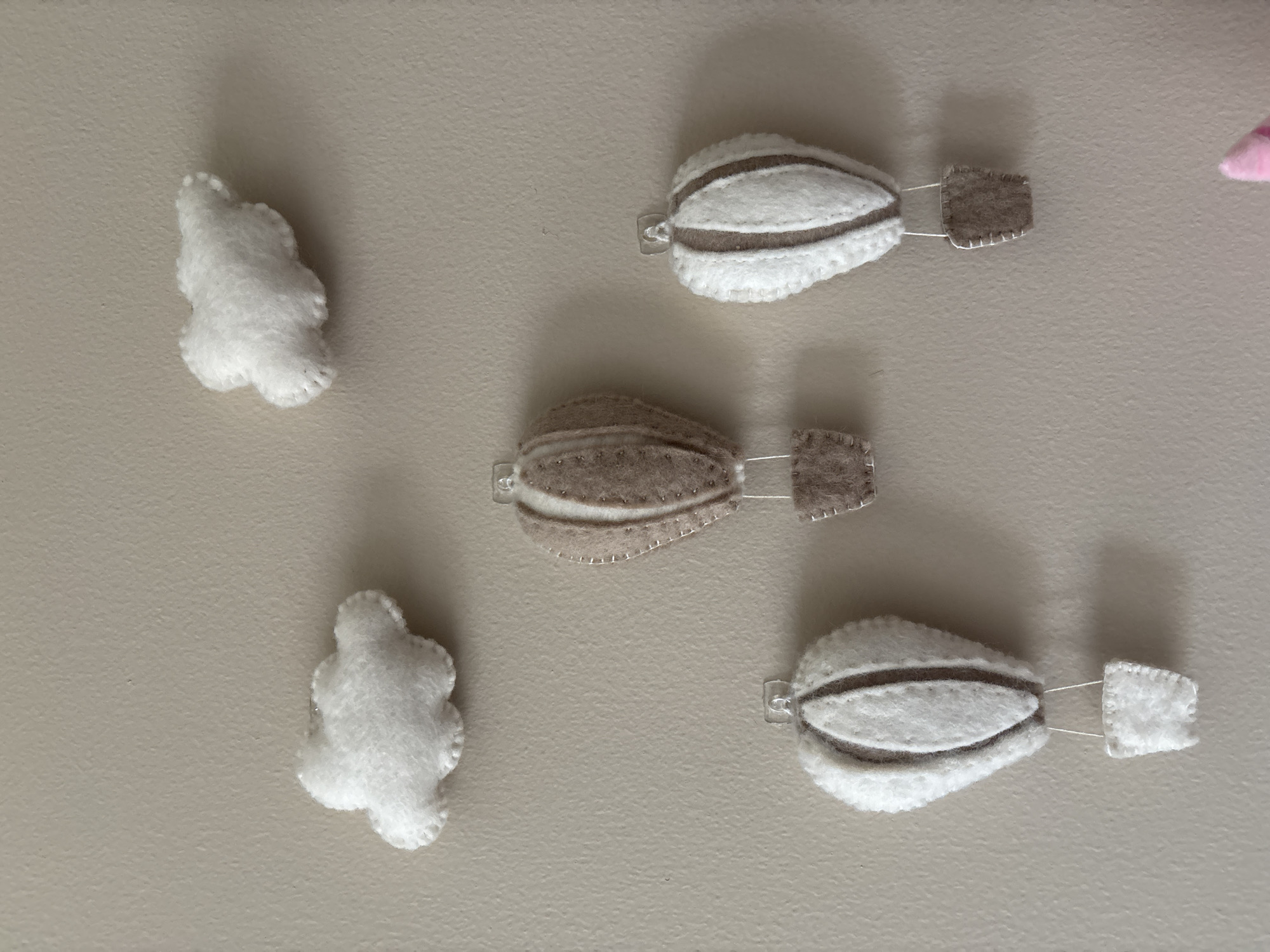 Three felt hot air balloons and two felt clouds are attached to a beige wall. The hot air balloons are white with brown stripes and small brown baskets. One hot air balloon is larger and positioned in the center of the image, while the other two are smaller and positioned on either side. The clouds are white and puffy. The image appears to be a close-up of a nursery or child's room decoration. The location in Dubai, United Arab Emirates is not determinable from the image.