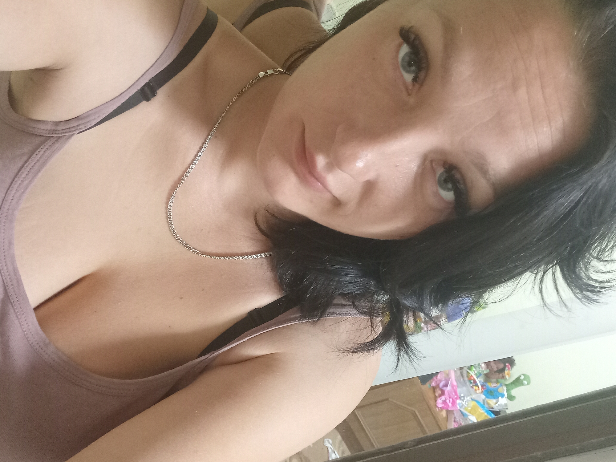 The image shows a close-up, selfie-style photograph of a woman with dark hair and light-colored eyes. She is wearing a light-colored tank top and a silver necklace. Her expression is neutral. The background appears to be an indoor setting, possibly a bedroom, with some blurred objects visible, including what seems to be a dresser and toys. There is no discernible indication of the specific location in Chuhuiv, Ukraine.
