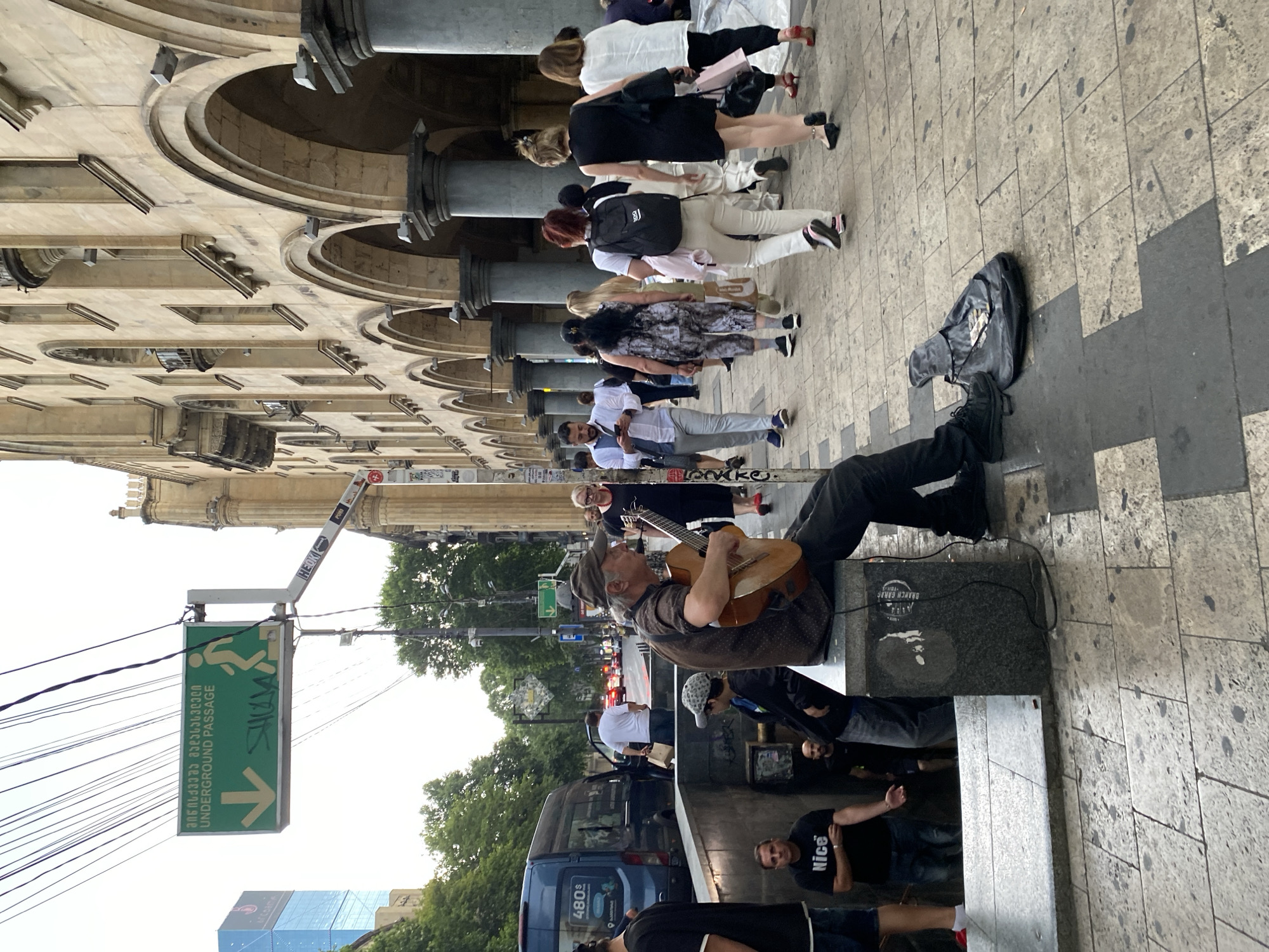 The image depicts a street scene in Tbilisi, Georgia, likely near an entrance to an underground passage. A man is seated on a stone ledge, playing an acoustic guitar. He wears a cap and a patterned shirt, with dark pants and shoes. Beside him, on the ground, is a black guitar case, open and containing what appears to be money. In the background, a blue van is parked on the left, and various pedestrians are walking along the sidewalk. A green sign with an arrow pointing downwards and text in Georgian and English indicating "UNDERGROUND PASSAGE" is visible, mounted on a pole above the street. The architecture of the building on the right features arches and columns, characteristic of older urban structures. The ground is paved with stone tiles.