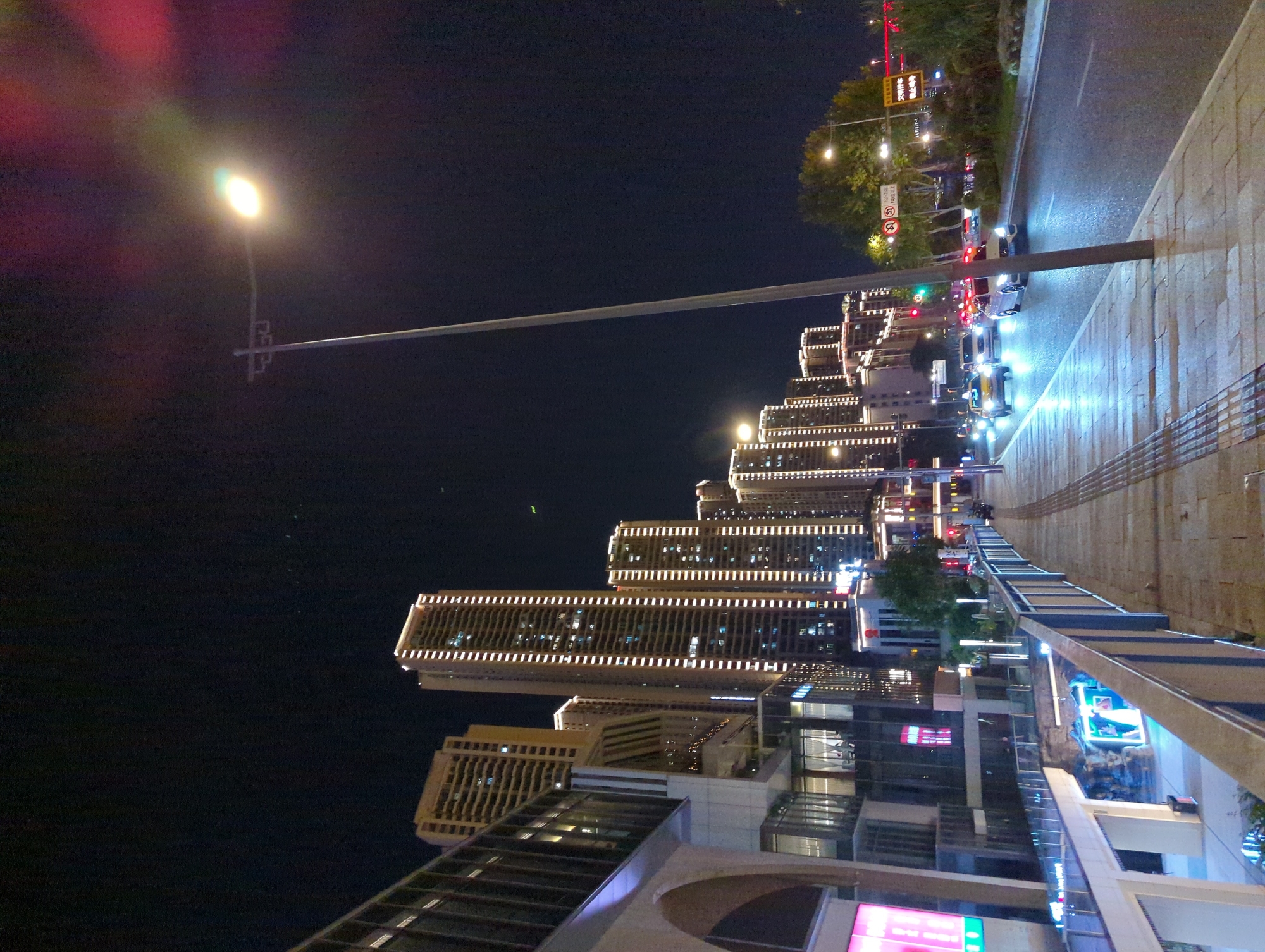 The image depicts a nighttime street scene in Jiangbeicheng Street, China, characterized by modern architecture and urban infrastructure. A wide sidewalk made of light-colored paving stones runs along the left side of the frame, bordered by a metal railing. Adjacent to the sidewalk is a multi-level building complex with extensive glass facades, illuminated from within. Further into the background, a cluster of tall, modern skyscrapers stands, their windows emitting a warm, yellow light, accentuating their verticality.
A street lamp with a bright, glowing bulb is prominently positioned in the center-right of the image, casting light on the surrounding area. The street itself is wet, reflecting the lights from vehicles and buildings. Several cars are visible on the road, including a white car in the foreground with its headlights on, and other vehicles further down the street. Traffic lights and street signs are also present, indicating an active urban environment. Lush green trees and vegetation line the right side of the road, contrasting with the concrete and glass structures. The sky above is dark, typical of nighttime, with a hint of light pollution.