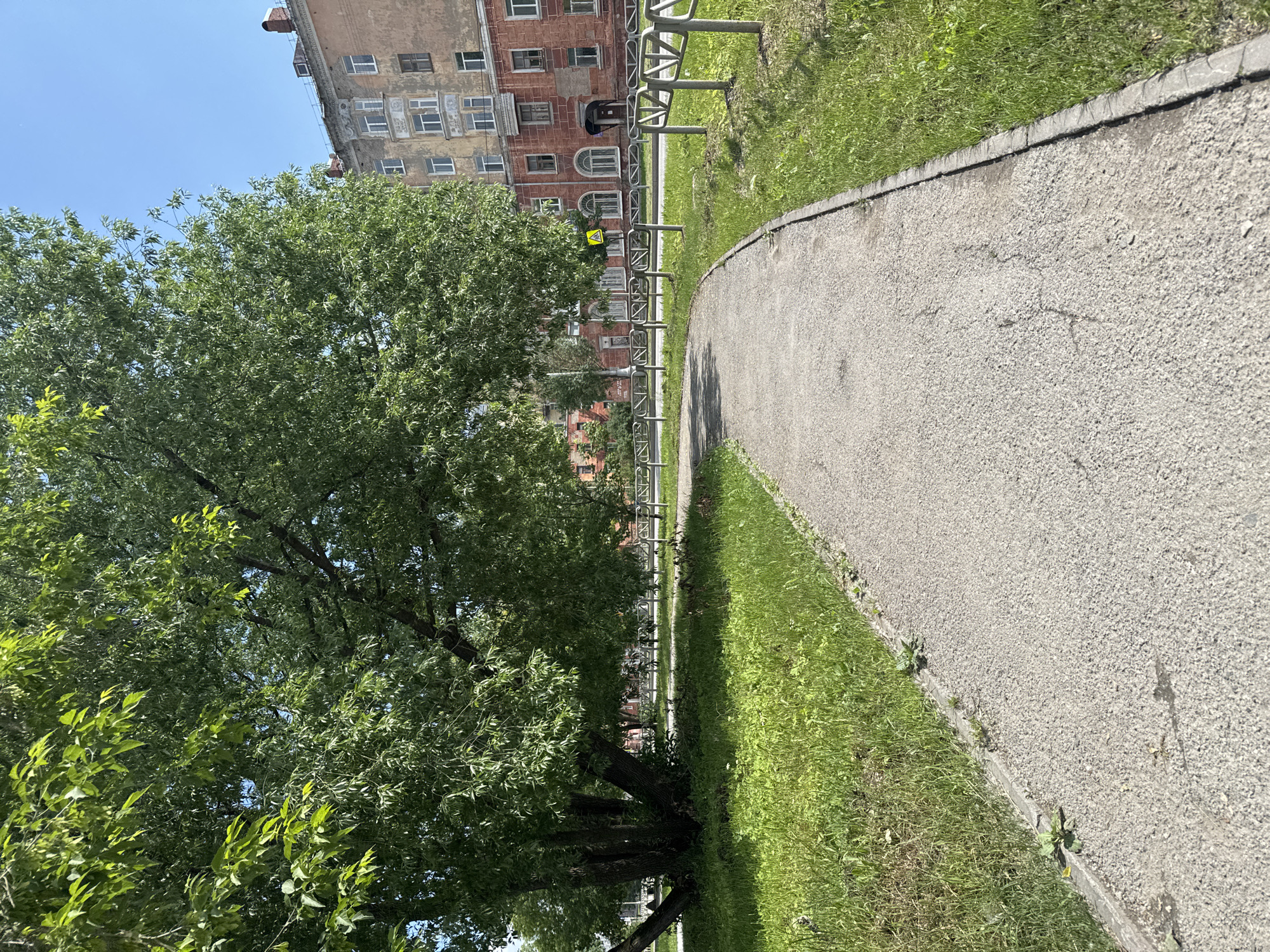 A paved pathway curves through a grassy area under the shade of a large, leafy tree. To the right of the path, a metal fence separates the green space from a sidewalk and a multi-story brick building with numerous windows. The building appears aged, with some visible signs of wear and tear. The sky above is clear and blue, indicating a sunny day. The scene is located in the city of Usolye, Russia, suggesting a typical urban park or residential area setting.