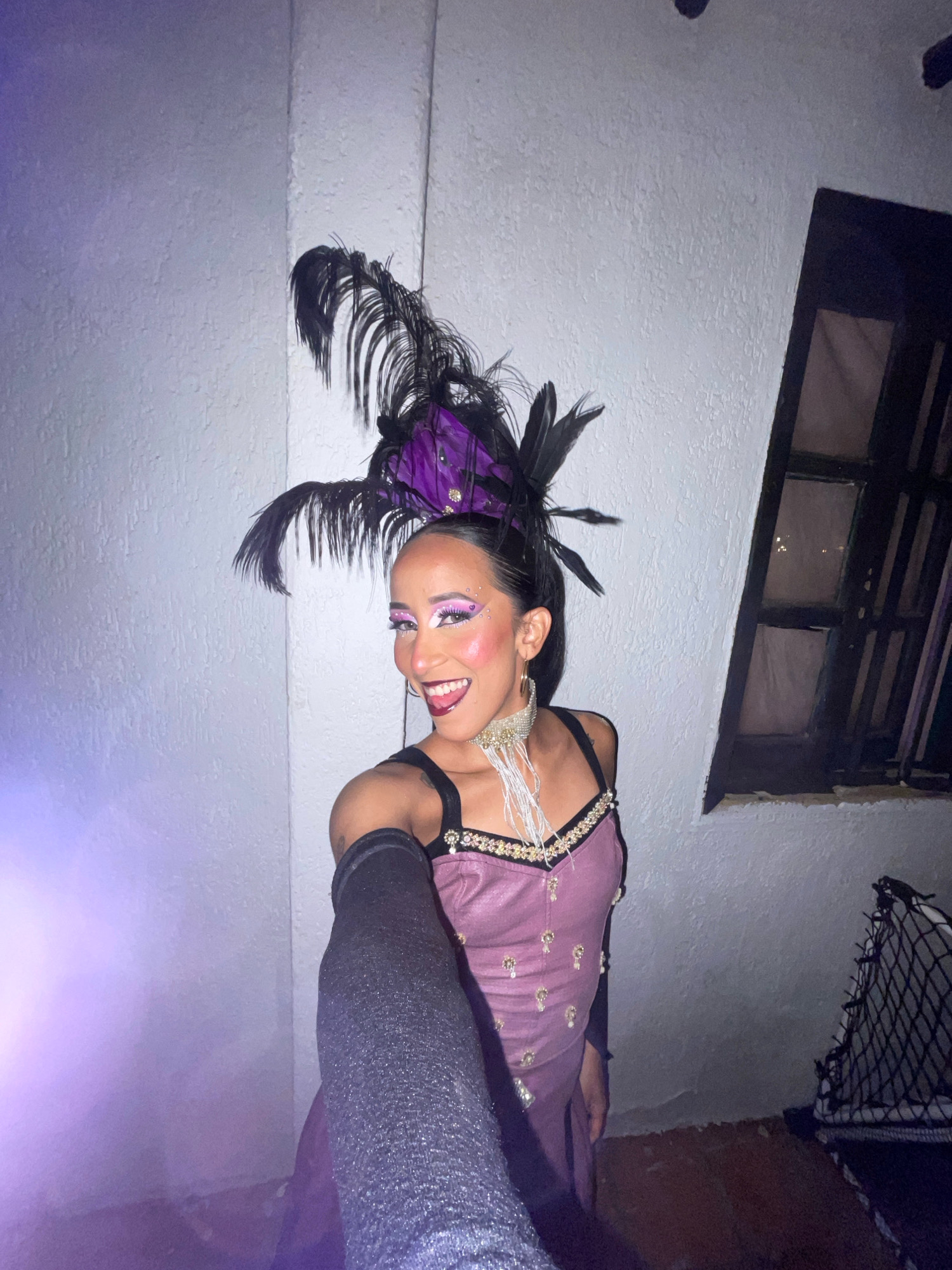 A woman in a purple and black feathered headdress, glittery eye makeup, and a sparkling purple costume stands in front of a textured white wall and a dark window. She wears long black sleeves and a decorative necklace. The lighting is dim, with a purple glow emanating from the left side of the frame. The floor appears to be rough concrete or dirt. In the background, a dark net-like object is visible. The scene suggests an outdoor or semi-outdoor urban setting, possibly at night, in Porlamar, Venezuela.