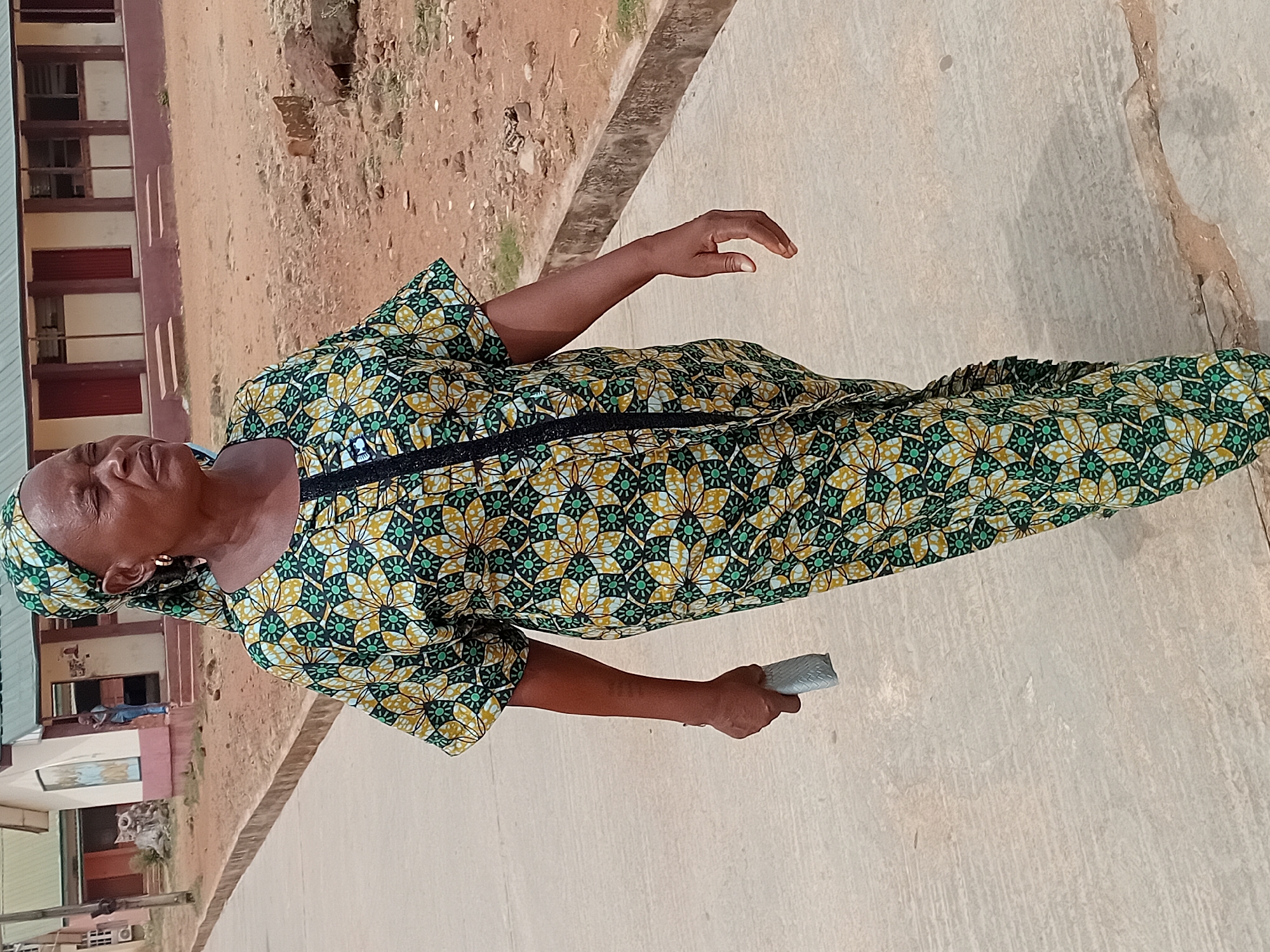 A woman stands on a sidewalk near a building, likely in Lokoja, Nigeria. She wears a patterned green and gold dress with a black trim. She has a headscarf matching the dress. In her right hand, she holds a small, gray object. The background includes a building with windows, brown soil, and a concrete sidewalk. The image is taken outdoors in daylight.