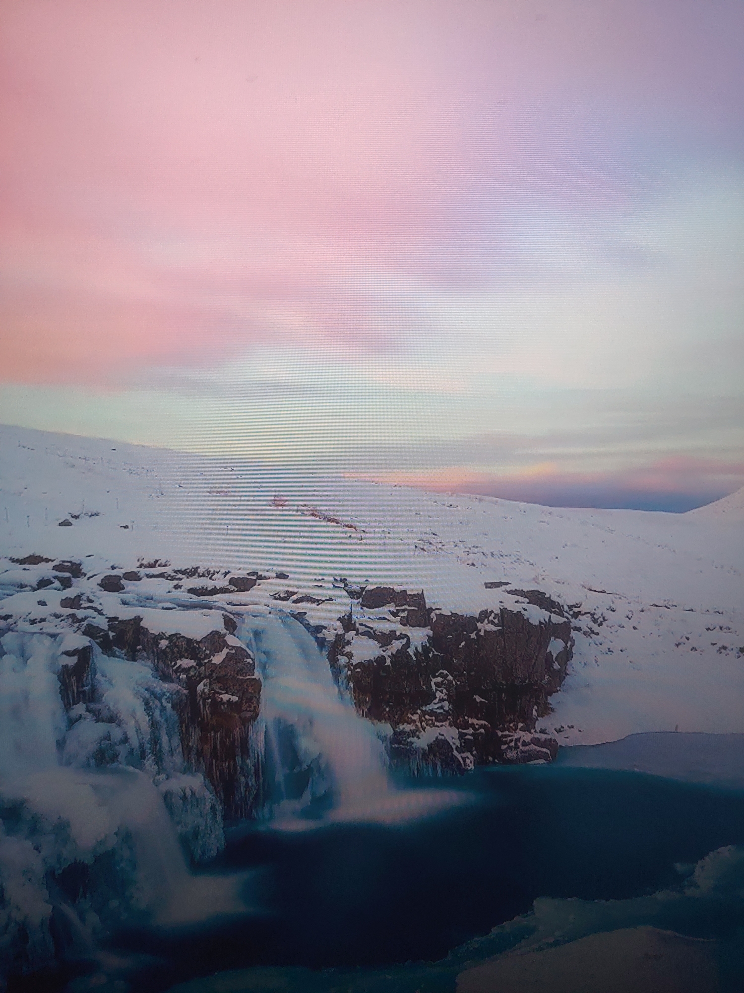 The image shows a waterfall cascading into a body of water against a snowy landscape. The sky above features a gradient of pink and white hues, suggesting sunrise or sunset. The surrounding terrain is covered in snow, with rocks and formations visible, some adorned with ice. The water in the pool at the base of the waterfall appears deep blue. The location appears to be in a cold climate, but precise coordinates or the city and country are Unknown.