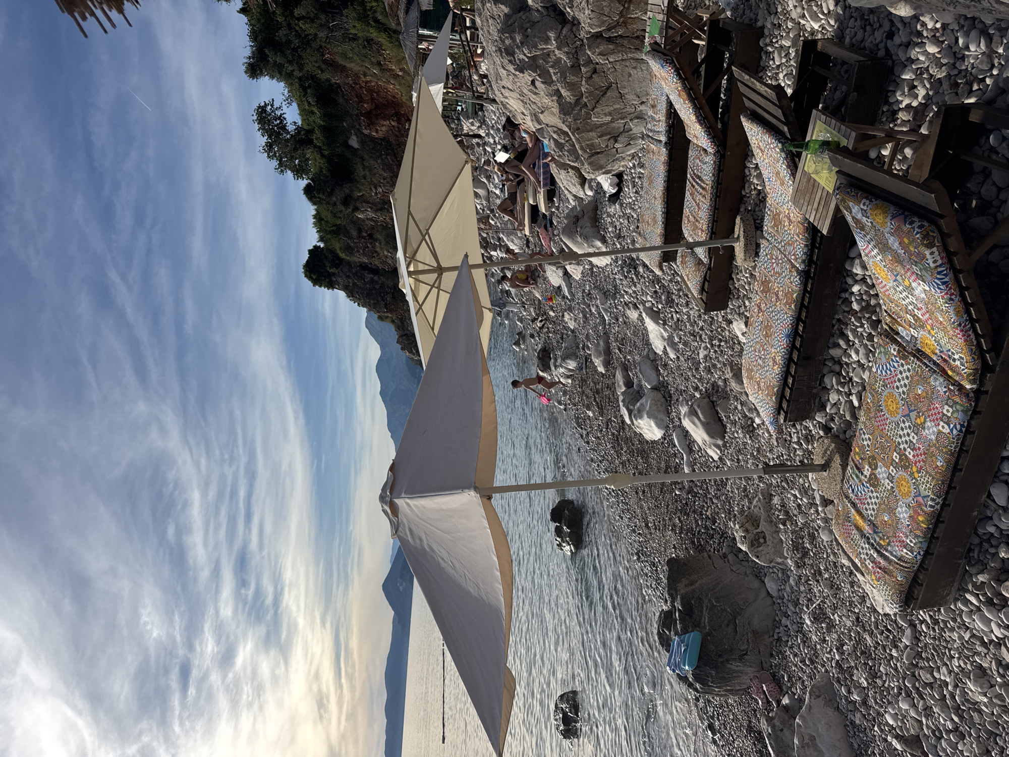 The image depicts a rocky beach in Rijeka Reževići, Montenegro, under a partly cloudy sky. Several beach chairs with colorful cushions are arranged on the pebble-strewn shore, some occupied by people sunbathing or relaxing. Two large, light-colored umbrellas provide shade. The calm sea stretches out towards a hazy horizon with mountains in the distance. A child with a pink toy is visible near the water's edge, and a green bottle sits on a small table next to one of the lounge chairs. The environment is natural, with rocky cliffs and vegetation visible on the right side of the frame.
