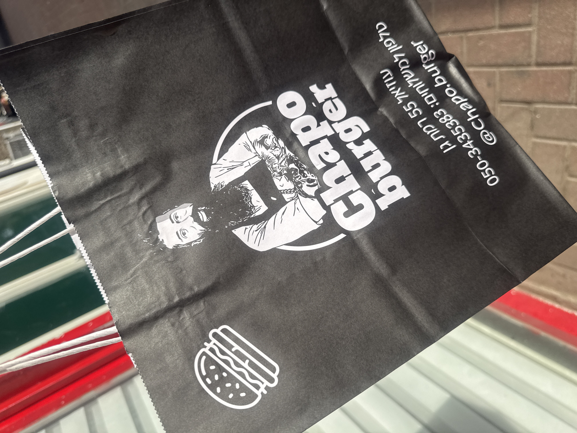 A black paper bag from "Chapo Burger," located at 55 Lanetz St, Ramat Gan, Israel, sits atop a surface with red, white, and grey stripes. The bag features a white cartoon image of a man's face above the words "Chapo Burger," along with a line-drawn burger illustration. Contact information, "050-3435383: ניוולמש אל דחיק" and "@chapo.burger," is printed in white.