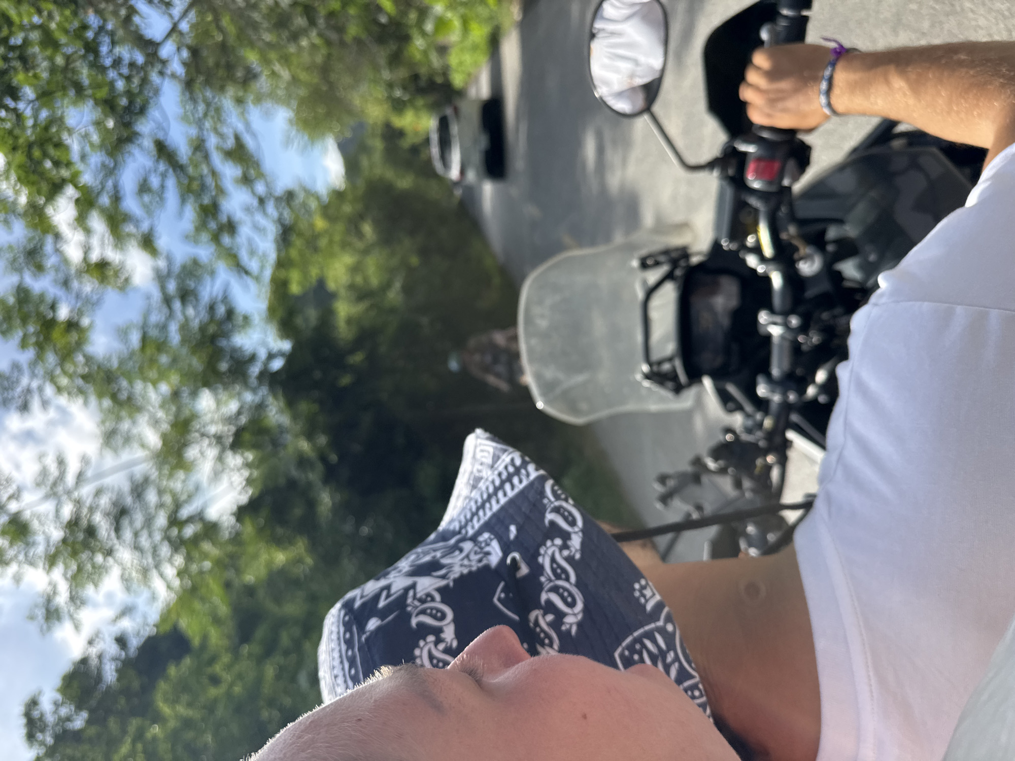 In Nam Hoa Lư Ward, Vietnam, a first-person perspective captures a ride on a motorcycle. The image focuses on the rider's arm and hand gripping the handlebar, adorned with a beaded bracelet. In the background, another motorcycle with a rider is visible, followed by a white car. The road is lined with lush green trees and foliage under a bright, partly cloudy sky, suggesting a rural or scenic route. The overall scene depicts travel and scenery in a Vietnamese locale.