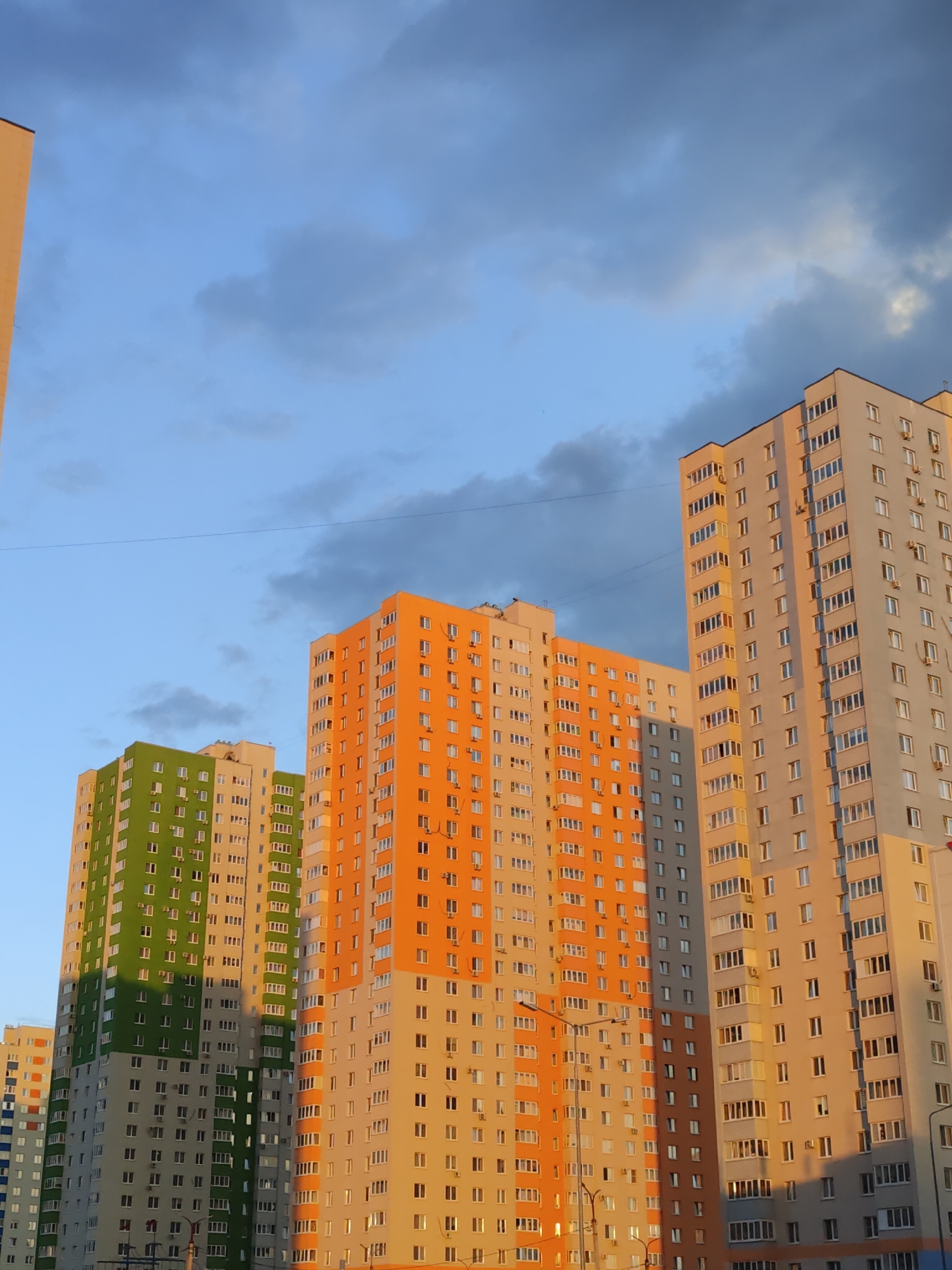 The image shows a row of modern, high-rise apartment buildings against a partly cloudy sky. The buildings vary in color, with one prominently featuring bright orange, another green, and a third a neutral beige or light gray. Many windows are visible on each facade, and some buildings have air conditioning units mounted outside. A street lamp is visible in the foreground, and a power line stretches across the sky. The overall impression is an urban residential area, possibly a neighborhood or district with newer construction.