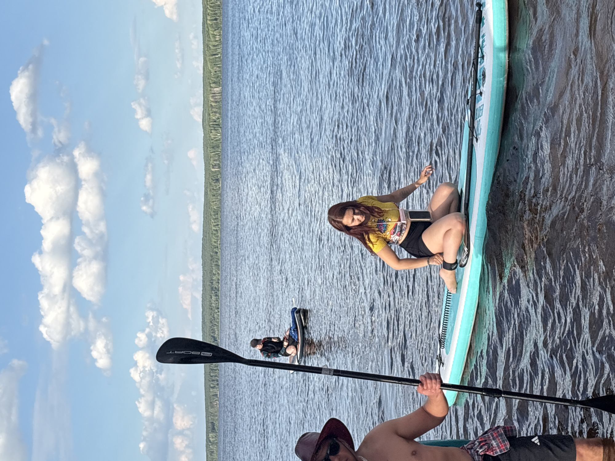 The image depicts a group of people engaged in paddleboarding on a body of water, likely a lake or river, with a tree-lined shore in the background under a partly cloudy sky. The perspective is from the water, looking towards the shore.
In the foreground, a person on the left, wearing a hat and sunglasses, is partially visible, holding a paddle. To the right, a woman with red hair is kneeling on a paddleboard, wearing a yellow shirt and black shorts. Another person is further back on a paddleboard, also kneeling.
The water is rippled, indicating some movement. The environment suggests a recreational outdoor setting. Based on the visual cues, this scene is consistent with locations like the Kama River embankment in Chaikovsky, Russia, known for its natural beauty and opportunities for water activities.