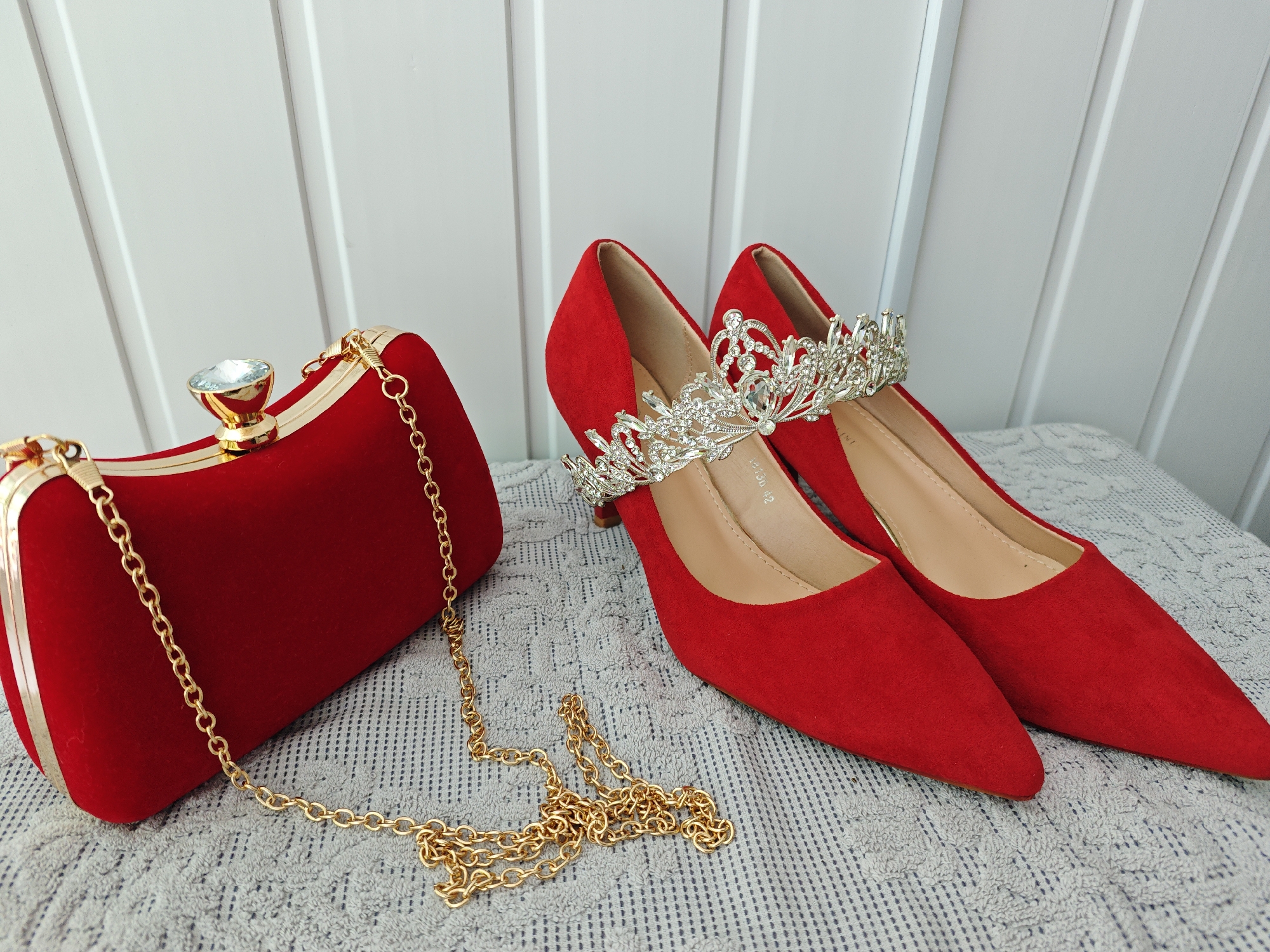 A still life photograph features a red clutch purse with a gold chain strap and a large crystal clasp, and a pair of red suede high-heeled shoes, each adorned with a silver tiara. The objects are arranged on a textured gray mat. The background consists of vertical white panels with grooves. The lighting is diffused and even. The photograph appears to be taken indoors. There is no information available in the image indicating that the image was taken in Markovskii, Russia.