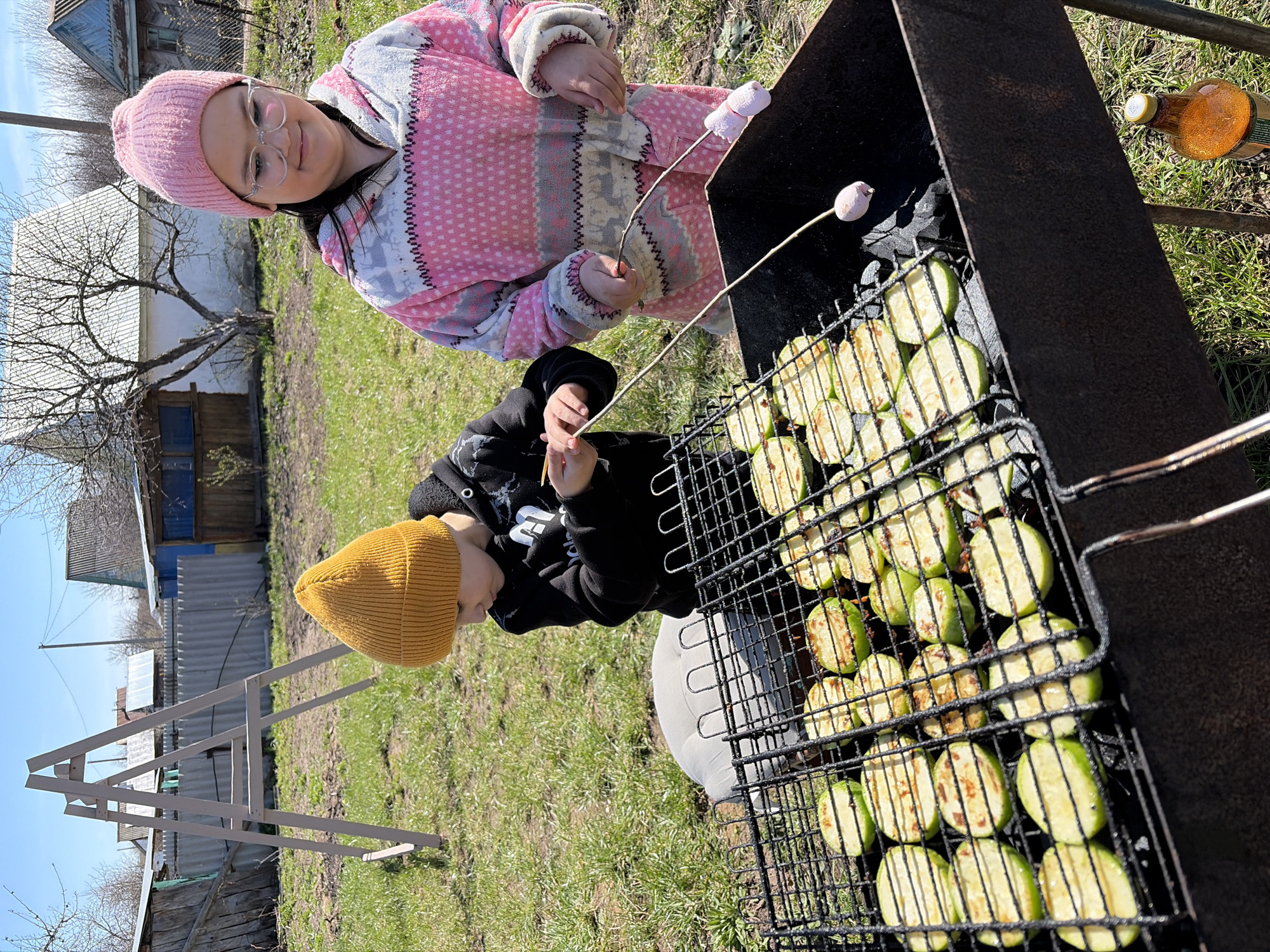 Two children stand near a barbecue grill in a grassy outdoor area in what appears to be the city of Neftekamsk, Russia. A girl with glasses, wearing a pink hat and pink patterned robe, holds a stick with a marshmallow on it. The second child, wearing a yellow beanie and a black jacket, holds a similar stick. The grill is filled with sliced zucchini. A bottle sits near the grill on the grass. In the background, there is a wooden structure and buildings typical of a suburban or rural area. The sky is clear and blue.