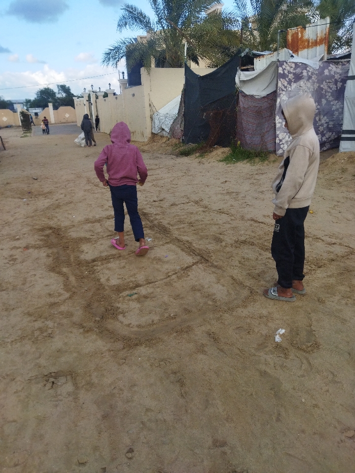 The image depicts an outdoor scene in the city of Khan Yunis, Palestinian Territory. The foreground consists of sandy ground, showing etched lines resembling a hopscotch grid. Two children are visible in the mid-ground. The child on the left wears a pink hooded sweatshirt, dark trousers, and pink sandals, facing away from the viewer. The child on the right wears a light beige hooded sweatshirt, dark trousers, and grey slide sandals, facing towards the left.
In the background, a sandy path extends into the distance, where several adult figures are walking, one appearing to carry a large white bag. To the right of the path, a light-colored wall with decorative pillars is visible, bordered by tall palm trees. Adjacent to the wall on the right, several makeshift shelters are constructed from various materials including dark tarps, patterned fabric, and corrugated metal sheets. The sky overhead is partially cloudy.