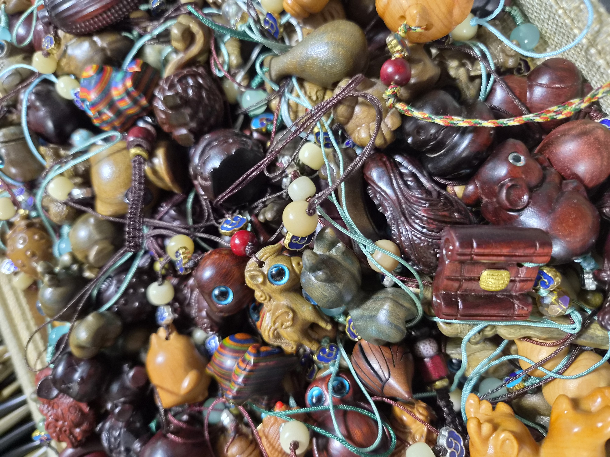 The image is a close-up, overhead shot of a large pile of intricately carved wooden charms and beads, all attached to various colored strings and cords. The objects are diverse in shape and color, featuring animal figures (such as horses and what appear to be mice or pigs), abstract shapes, and what looks like a miniature book or scroll. Many of the carvings have bright blue eyes. The overall impression is of a jumbled collection of handmade crafts. There is no visible context regarding the city of Qinhuangdao, China, in the image itself. The scene is densely packed with these decorative items.