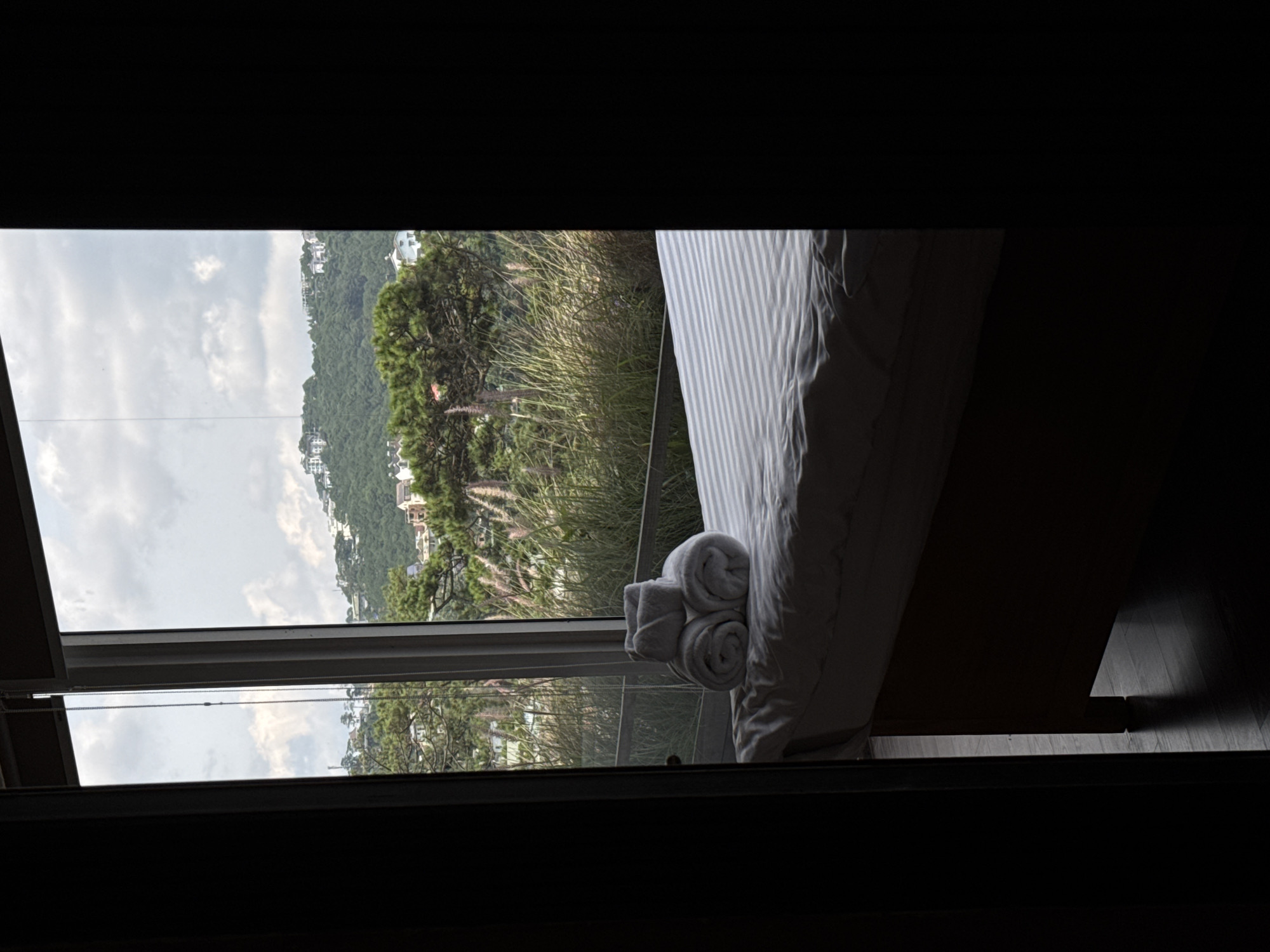 The image depicts a room with a bed covered in a striped duvet, positioned in front of a large window. On the bed, there are three neatly folded white towels arranged in a stack. Through the window, a lush green hillside is visible, dotted with houses and trees. The sky is overcast. The scene is framed by dark doorways on either side, suggesting the room is viewed from a hallway or another interior space. The overall impression is one of a serene and comfortable lodging, likely in a location with a scenic natural backdrop, such as Da Lat, Vietnam, known for its hilly terrain and European-inspired architecture.