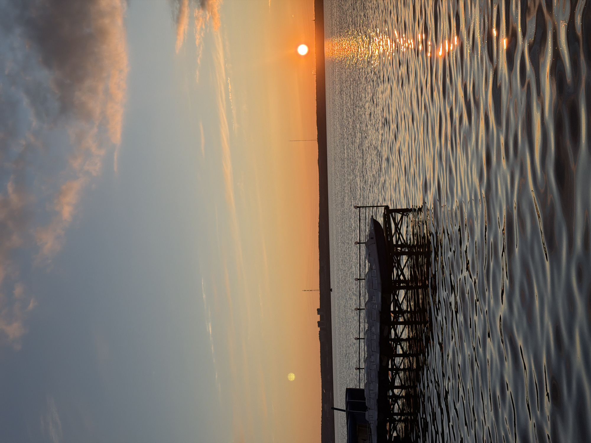 A serene landscape photograph captures a sunset or sunrise over a large body of water, likely the Izhevsk Pond in Izhevsk, Russia. The sun, a bright orange-yellow orb, is positioned just above the distant horizon, casting a warm, golden glow across the sky and water.
The sky occupies the upper portion of the frame, transitioning from a soft orange and yellow near the sun to a lighter blue-grey further up and to the left. Wispy, elongated clouds, tinged with pink and orange from the low sun, are scattered across the left side of the sky.
The horizon line, visible across the middle of the image, features a dark, silhouetted landmass, possibly a distant cityscape, with a tall, slender antenna or tower standing prominently.
The lower half of the image is dominated by the water, which is covered in distinct ripples. The sun's intense light creates a shimmering, sparkling path of reflection that stretches across the water's surface from the horizon towards the viewer. The water appears dark grey-blue in shadowed areas and takes on a golden hue where the light reflects directly.
In the foreground, on the right side, a dark, metallic-looking structure, possibly part of a pier or dock, extends into the water. Its crisscrossing beams and supports are silhouetted against the bright water. Small white markers or buoys are visible along the structure. Part of a dark boat or watercraft is also partially visible in the bottom right corner, next to the pier.
The scene conveys a peaceful and tranquil atmosphere, devoid of human activity. No visible text is present in the image.