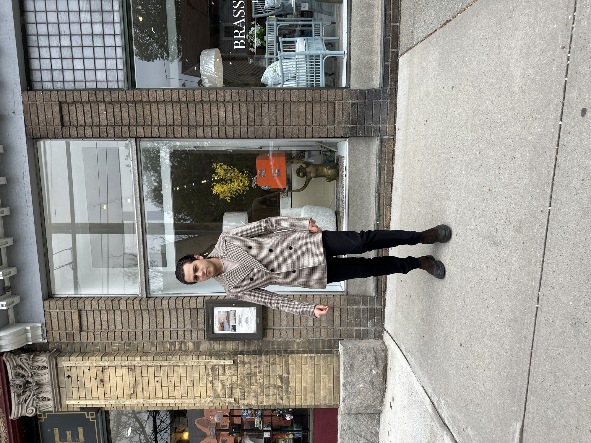 A man stands upright on a concrete sidewalk in front of a brick building featuring several storefronts. He is centrally positioned, facing the camera with a neutral expression. He wears a double-breasted jacket with a brown and white houndstooth or plaid pattern, a light-colored top underneath, black trousers, and dark boots. His dark hair is neatly styled.
The background consists of a multi-story building facade. The lower part is primarily light brown brickwork, while the upper section has large display windows. Through the window to the man's right, a sign partially reads "BRASS," and inside, light-colored rattan furniture is visible. The window to his left displays various decorative items, including a golden monkey statue, an orange box, a vase with yellow flowers, and a white lampshade. Another window section features a grid-like pattern. Further down the facade, a framed notice is attached to the brickwork, though its text is illegible. The sidewalk is plain concrete, with visible expansion joints.
The scene appears to be set during daytime, with soft, diffused lighting suggesting an overcast sky. The man is posing for a photograph. The overall setting indicates a commercial street or town center in the Village of Cold Spring, USA.