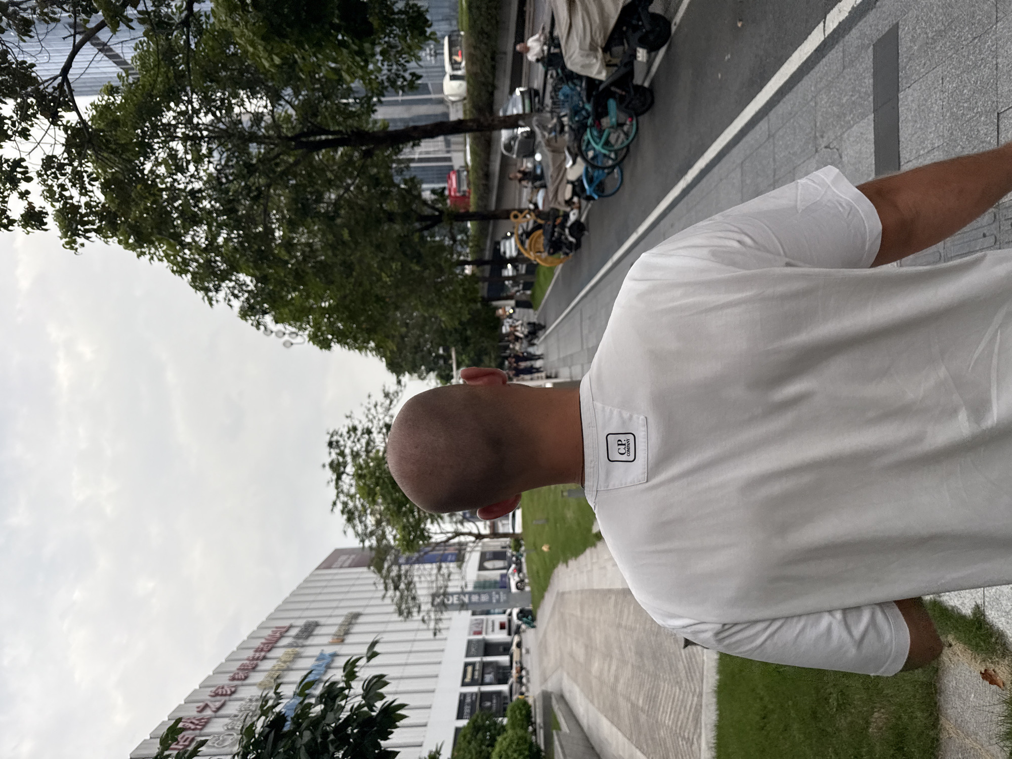 A man with a shaved head walks away from the camera down a paved sidewalk. He is wearing a white t-shirt with a small black and white logo on the back, just below the collar. To his left is a green lawn and a modern, multi-story building with a white facade and vertical lines. The building has signs with text in both Chinese and English, including "GRANDVIEW MALL". To his right, across a narrow strip of grass, is a road with several vehicles, including motorcycles, scooters, and cars. In the background, there are trees and more modern buildings. The sky is overcast.