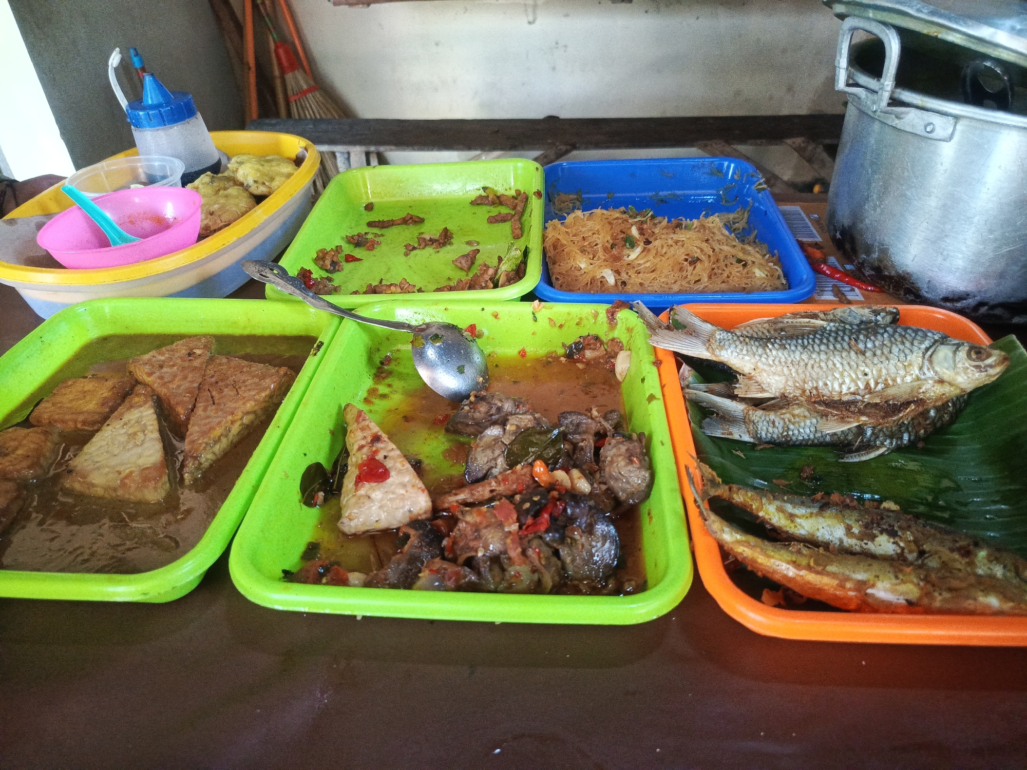 This image depicts a close-up view of various Indonesian dishes displayed in plastic trays on a brown surface, likely a table. In the foreground, several green plastic trays hold different food items. The leftmost green tray contains pieces of tempeh submerged in a brown sauce. The adjacent green tray has a triangular piece of tempeh and what appears to be pieces of meat or offal in a reddish-brown sauce with chili. To the right, an orange tray holds two fried small fish in the foreground and a larger fried fish on top of a green banana leaf in the background. Behind these, a blue tray contains fried vermicelli noodles. In the upper left, a yellow oval container holds fried fritters, a pink bowl with a turquoise spoon, and a blue-capped squeeze bottle containing a dark liquid. A large metal pot is visible in the upper right background, partially obscuring a broom and dustpan. The scene appears to be set up for self-service or a food stall. The overall lighting suggests an indoor setting, possibly a food court or a casual eatery. There is no indication of a specific location within the city of Semarang, Indonesia, within the image itself.
