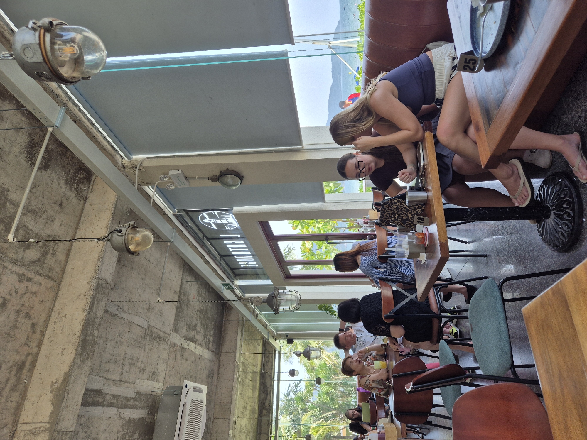 The image depicts the interior of a restaurant or cafe in Da Nang, Vietnam, with a view of the sea and lush tropical vegetation through large windows. The architectural style features an exposed concrete ceiling, industrial-style pendant lights, and large glass windows that offer a panoramic view of the outdoors.
Several groups of people are seated at tables, engaged in conversations and dining. In the foreground, two young women are looking at a laptop at a wooden table. To their left, a couple is seated at a table, with the man appearing to be telling a story and laughing. Another woman is seated at the same table, looking towards the man. Further to the left, another group of people are dining, with one woman wearing glasses and another with short dark hair.
The overall atmosphere is casual and relaxed, with natural light flooding the space. The presence of the sea and tropical plants suggests a coastal location within Da Nang. The decor includes simple wooden furniture, a leather banquette, and some decorative elements like the industrial pendant lights.