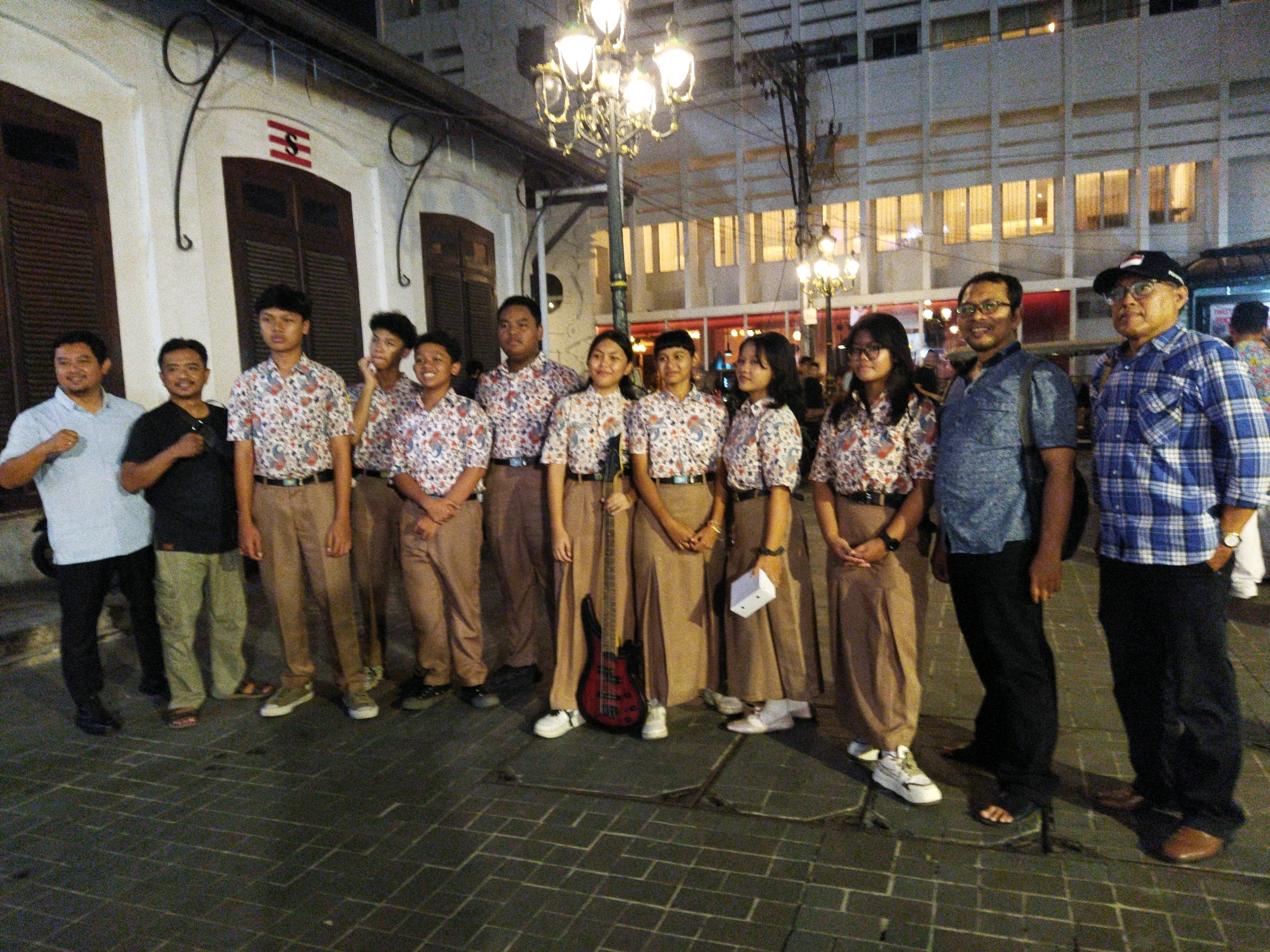 The image depicts a group of ten people, six males and four females, standing on a brick-paved sidewalk at night. The group appears to be posing for a photograph. The majority of the individuals, specifically the six young people in the center, are wearing matching white shirts with a colorful, repeating floral or paisley pattern and light brown pants or skirts. A red electric guitar is standing upright between two of the young people. Behind the group, there is a building with white walls and dark shutters on the ground floor, and a modern building with multiple illuminated windows on the upper floors. A street lamp with ornate fixtures is positioned behind the group. The people are of varying ages and ethnicities, with most appearing to be of Southeast Asian descent. The setting suggests a street scene in a city, possibly a historical district given the architecture of the building on the left. The overall lighting indicates it is nighttime, with artificial lights from the street lamp and buildings illuminating the scene. This scene is consistent with a public space in the Kota Lama area of Semarang, Indonesia, known for its colonial-era architecture and cobblestone streets.