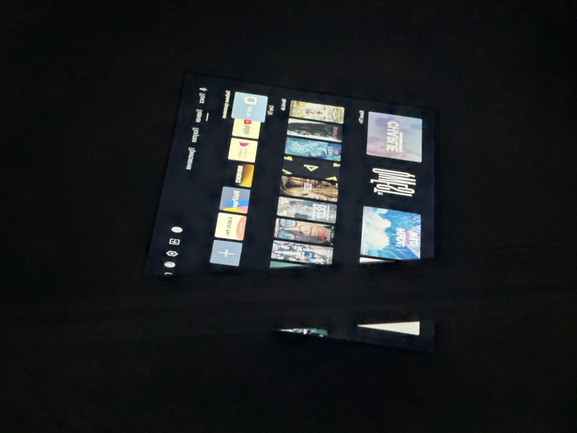The image shows a screen displaying a user interface with multiple tiles, likely for a streaming service or smart TV. The interface is in a horizontal orientation, and the tiles appear to be a mix of application icons and content previews, possibly movies or TV shows. Some of the text on the tiles is blurred or appears to be in a reversed or mirrored font, suggesting it may be a reflection or a display issue.
The screen is positioned against a dark background, which could be a wall or a darkened room. The lighting is dim, with the screen being the primary source of illumination. There are no people or animals visible in the image.
Regarding the location in Saratov, Russia, there is no visible information within the image itself to suggest this location. The image focuses solely on the screen content and its immediate surroundings, which are generic and lack any identifying landmarks or contextual clues specific to Saratov.