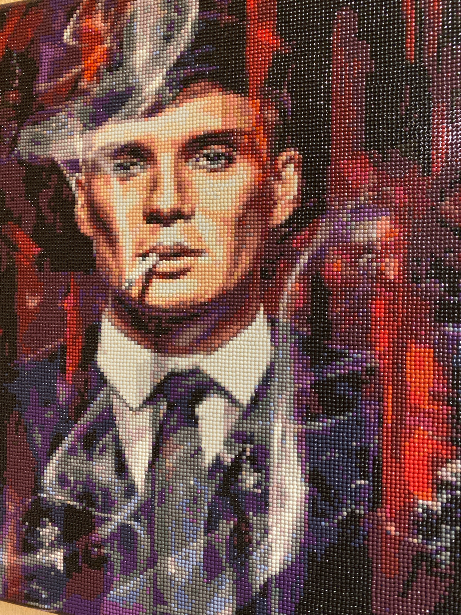The image is a diamond painting of a man in a suit and hat, smoking a cigarette. The man has light skin and is looking to his right. He is wearing a white shirt and a patterned tie and suit. The background is a mix of red, purple, and black colors. The diamond painting is likely a close-up of the man's face and upper body.
There is no information in the image to suggest that it is located in the city of Korablino, Russia, or any other specific geographic location. The scene is entirely contained within the artwork itself.