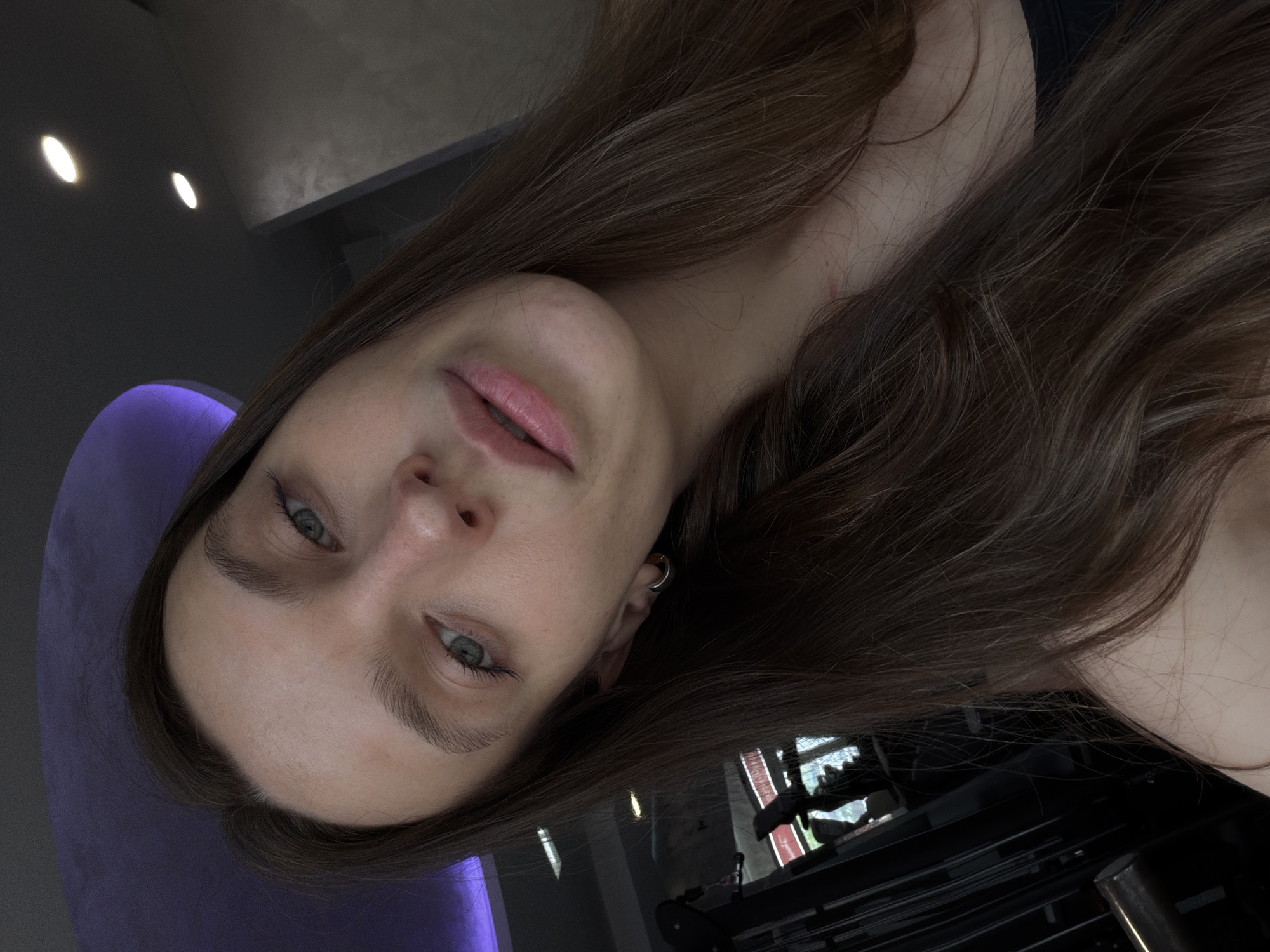 A woman with long brown hair is pictured in an upside-down selfie. Her face is centered, with her eyes gazing toward the viewer. She is wearing a small hoop earring in her left ear. Behind her and to the left is a purple chair. In the background, a glimpse of a fitness machine suggests she might be in a gym. Based on the lighting and equipment, she appears to be indoors, possibly in Budva, Montenegro if the location information is accurate. However, there are no elements in the image to confirm that information.
